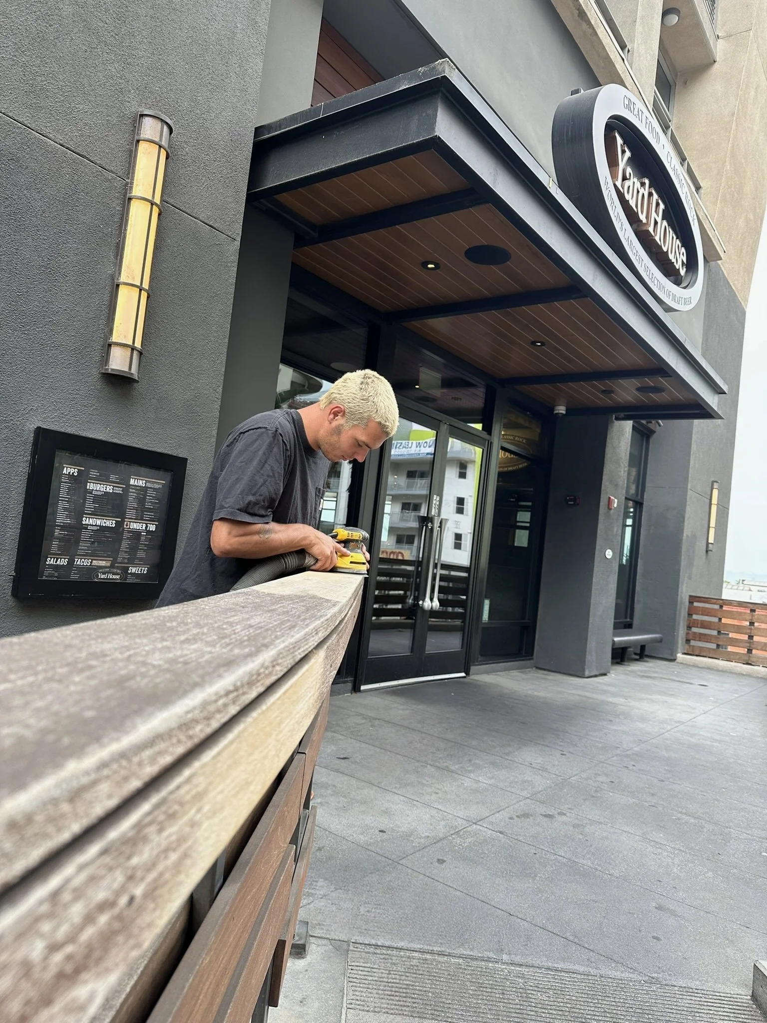 Man sanding a wooden railing outside Yard House restaurant.
