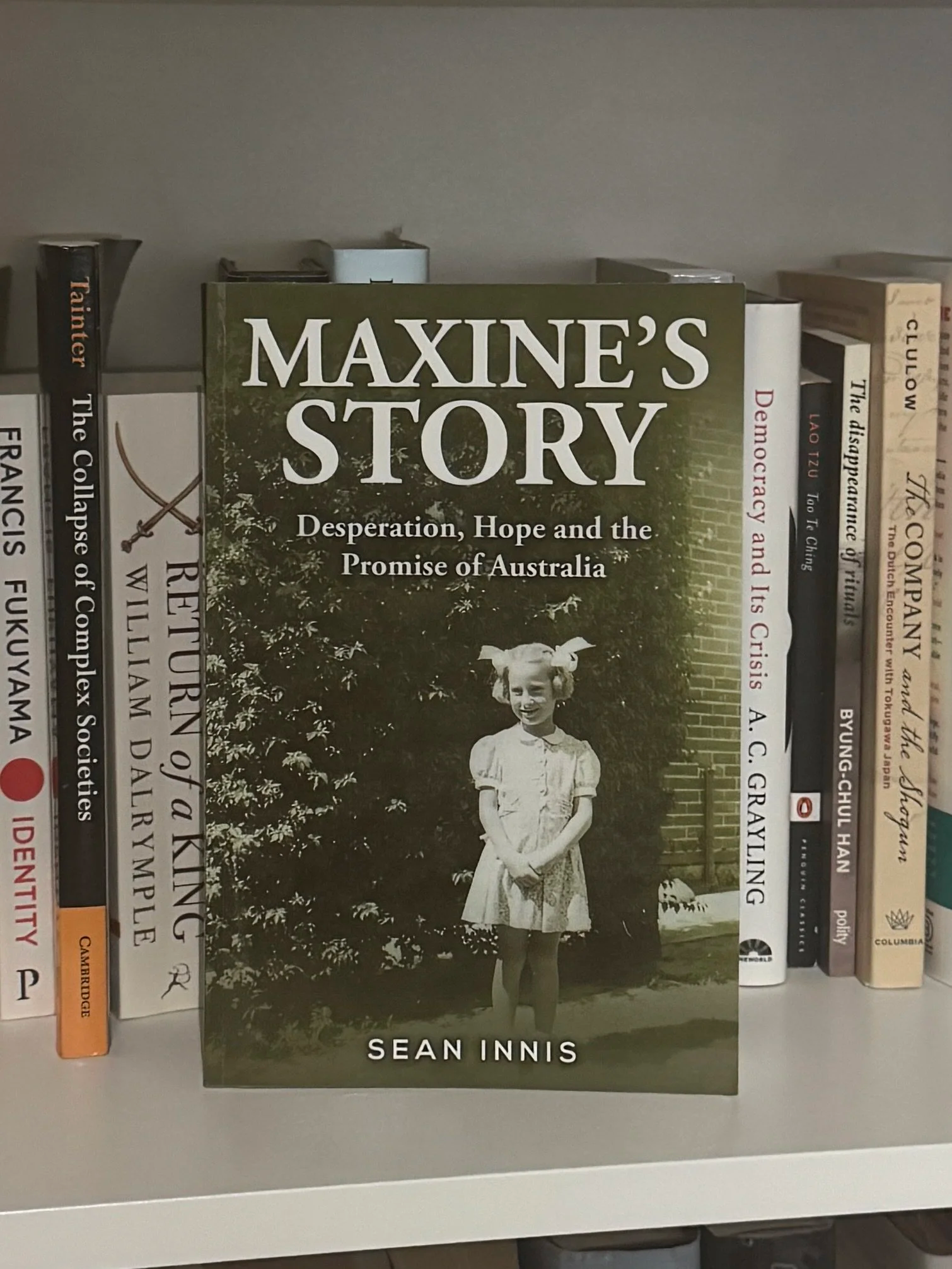 Book titled 'Maxine's Story: Desperation, Hope and the Promise of Australia' by Sean Innis, featuring a black and white photograph of a young girl in a dress, standing outdoors in front of a bush, on a bookshelf with other books.