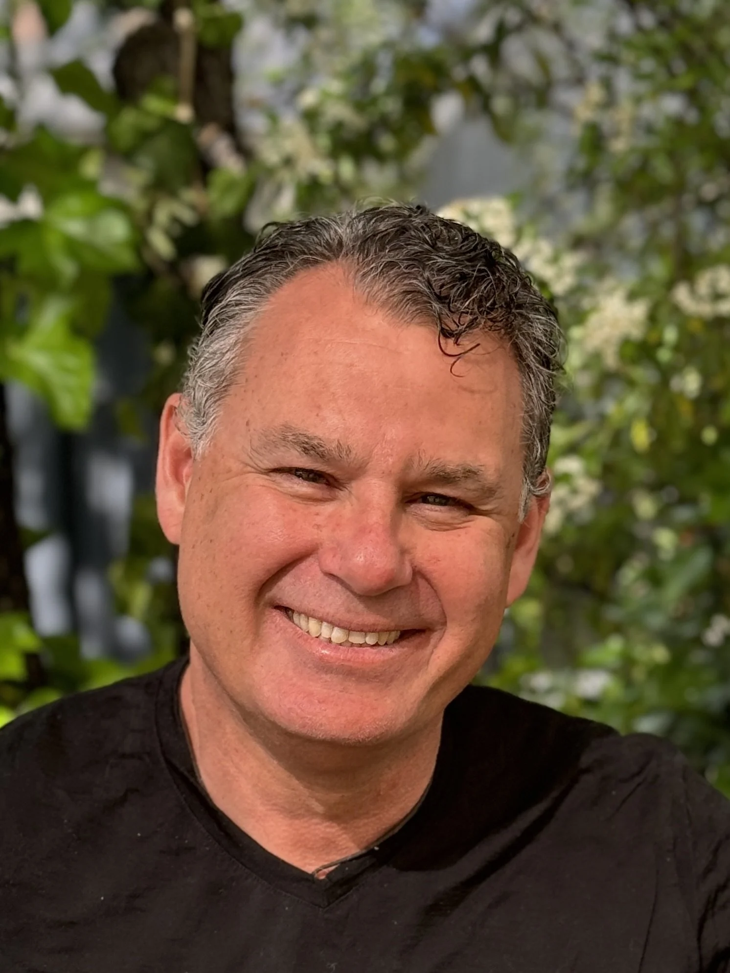Close-up photo of a smiling man with dark, slightly curly hair, wearing a black shirt, outdoors with green foliage in the background.