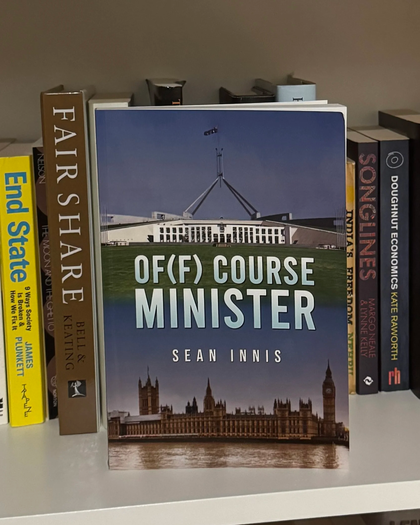 Book titled "OF (F) COURSE MINISTER" by Sean Innis on a shelf surrounded by other books, with the Australian Parliament House in the background on the cover.