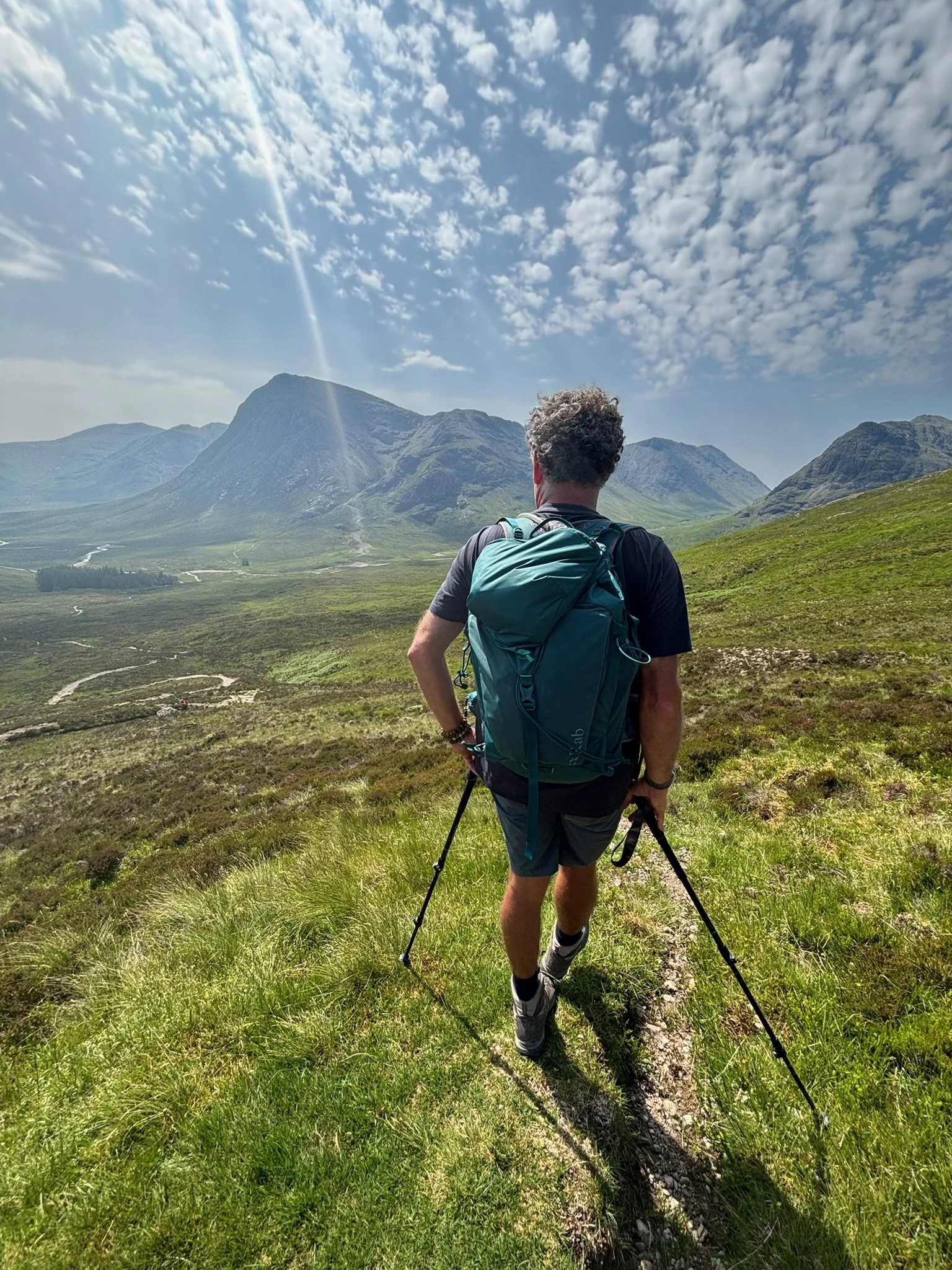 A man with a backpack and trekking poles walking on a narrow trail in a green mountainous landscape under a partly cloudy sky.