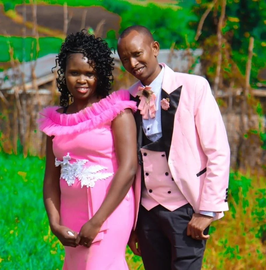 A man and woman dressed formally, standing outdoors with greenery and trees in the background. The woman wears a pink dress with ruffled shoulders and white floral embroidery, while the man wears a pink and black tuxedo with a pink bow tie.