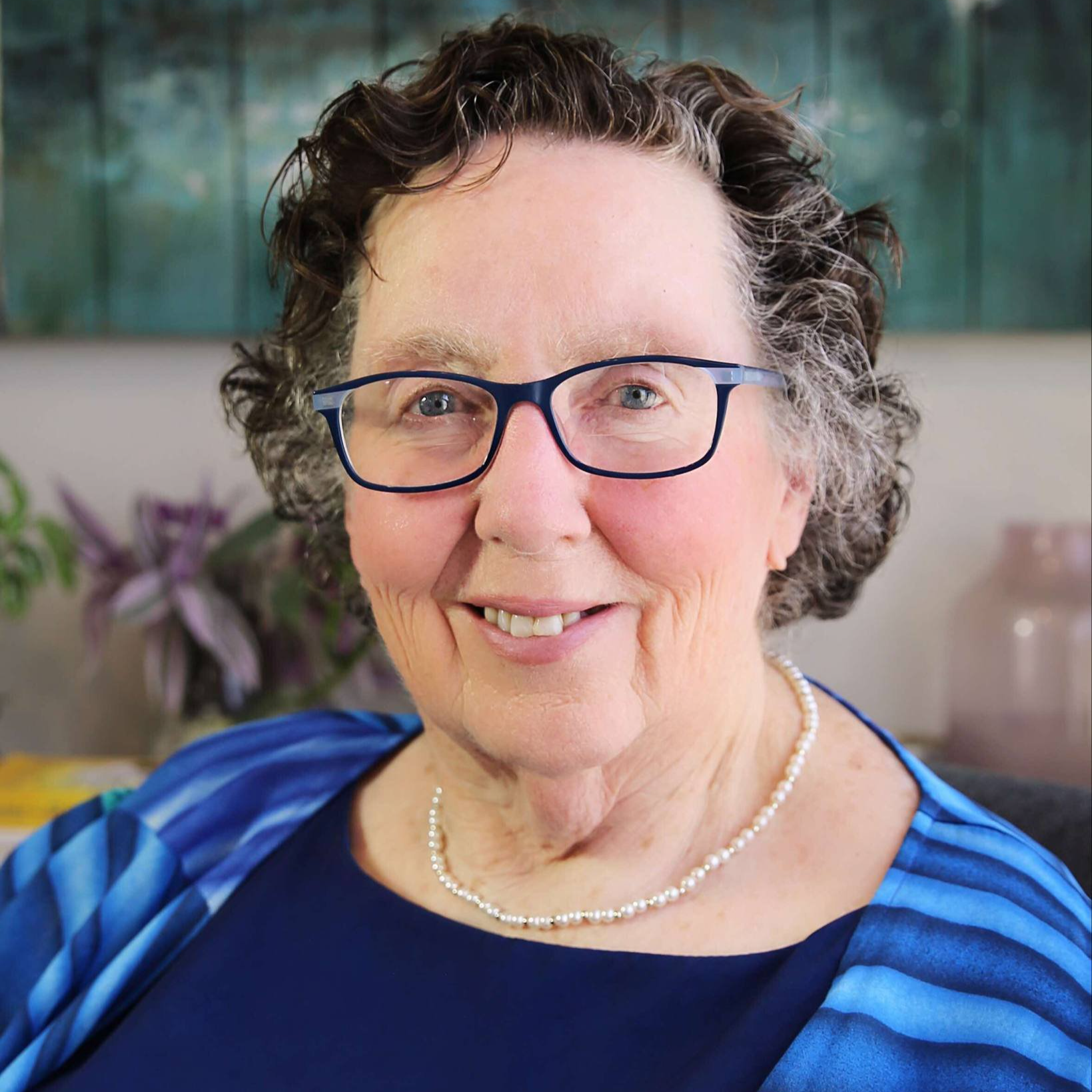 A smiling older woman with short, curly gray hair, wearing blue glasses, a pearl necklace, and a blue outfit, sitting indoors with a blurred background of plants and artwork.