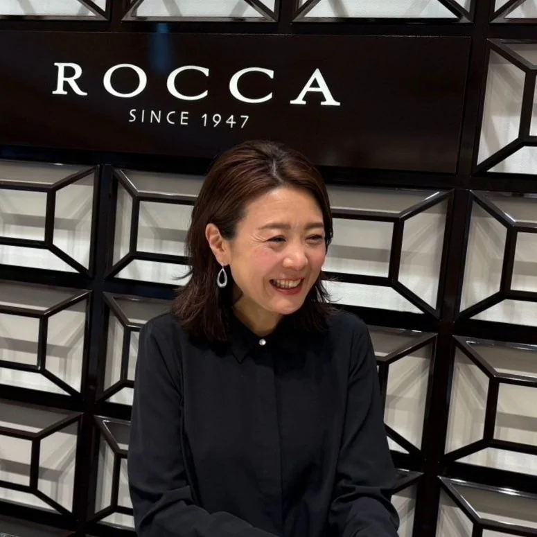 笑顔の女性が黒いシャツを着て、アートな壁の前に立っている写真。背景には『ROCCA SINCE 1947』と書かれたサインがある。