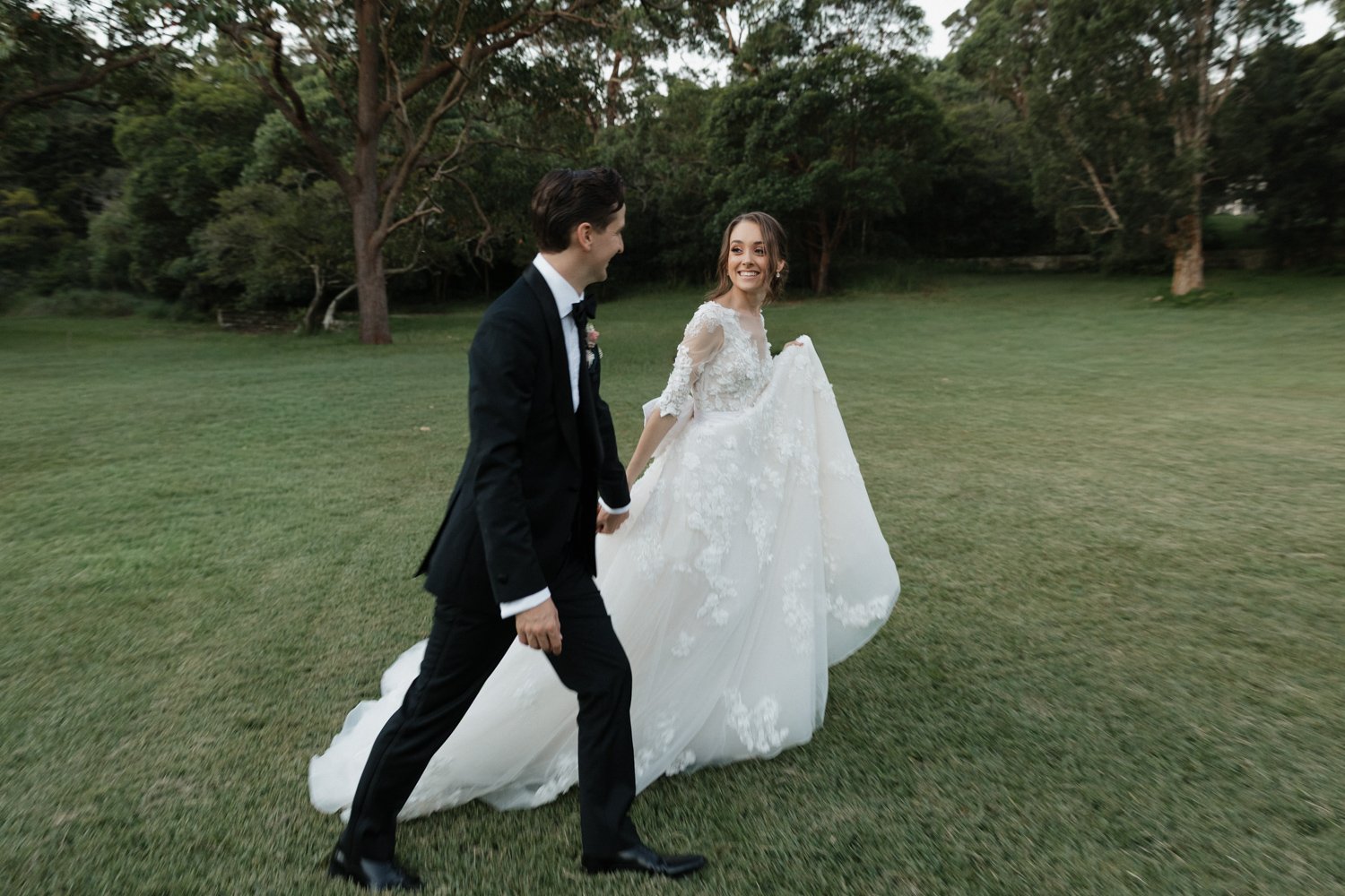 Bride and groom walking hand in hand across a grassy lawn surrounded by tall trees. The bride wears a flowing lace wedding gown with sheer sleeves and smiles at the groom, who is dressed in a black tuxedo. The scene feels natural and candid.