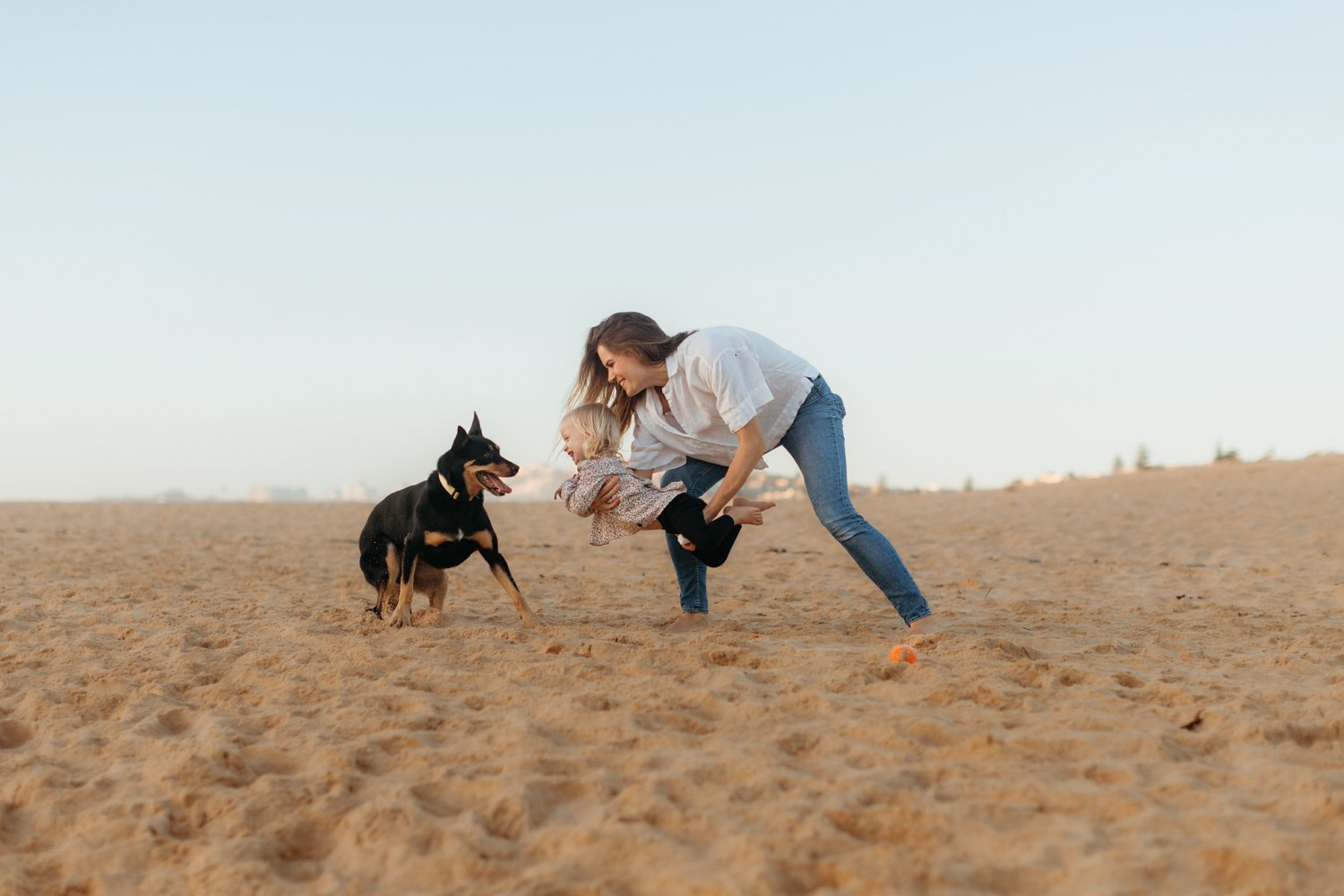 a mum holding her toddler and playing with their black and tan kelpie on a sandy beach