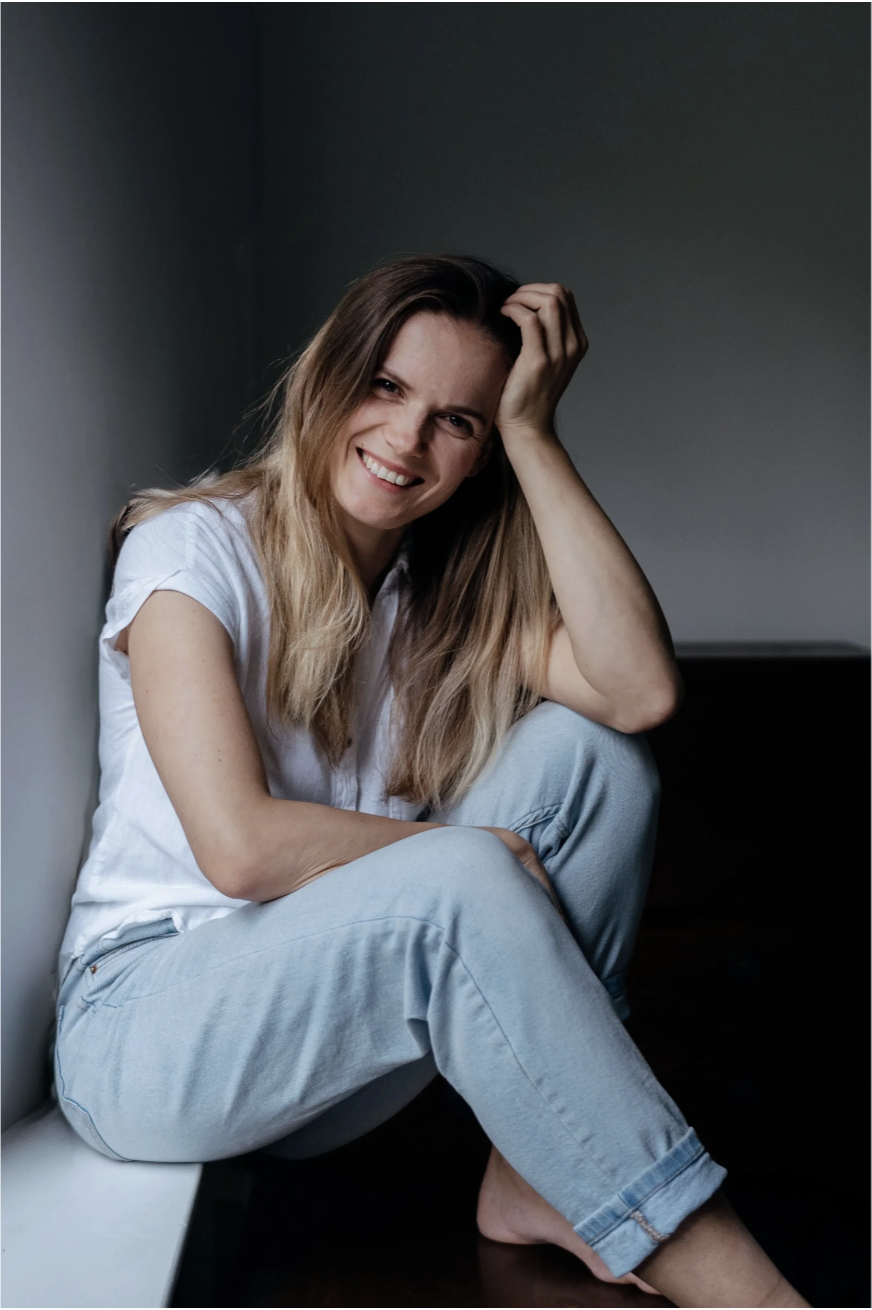This is the photographer. A woman with long brown hair smiling warmly, sitting casually in a white top and jeans, resting her head on her hand