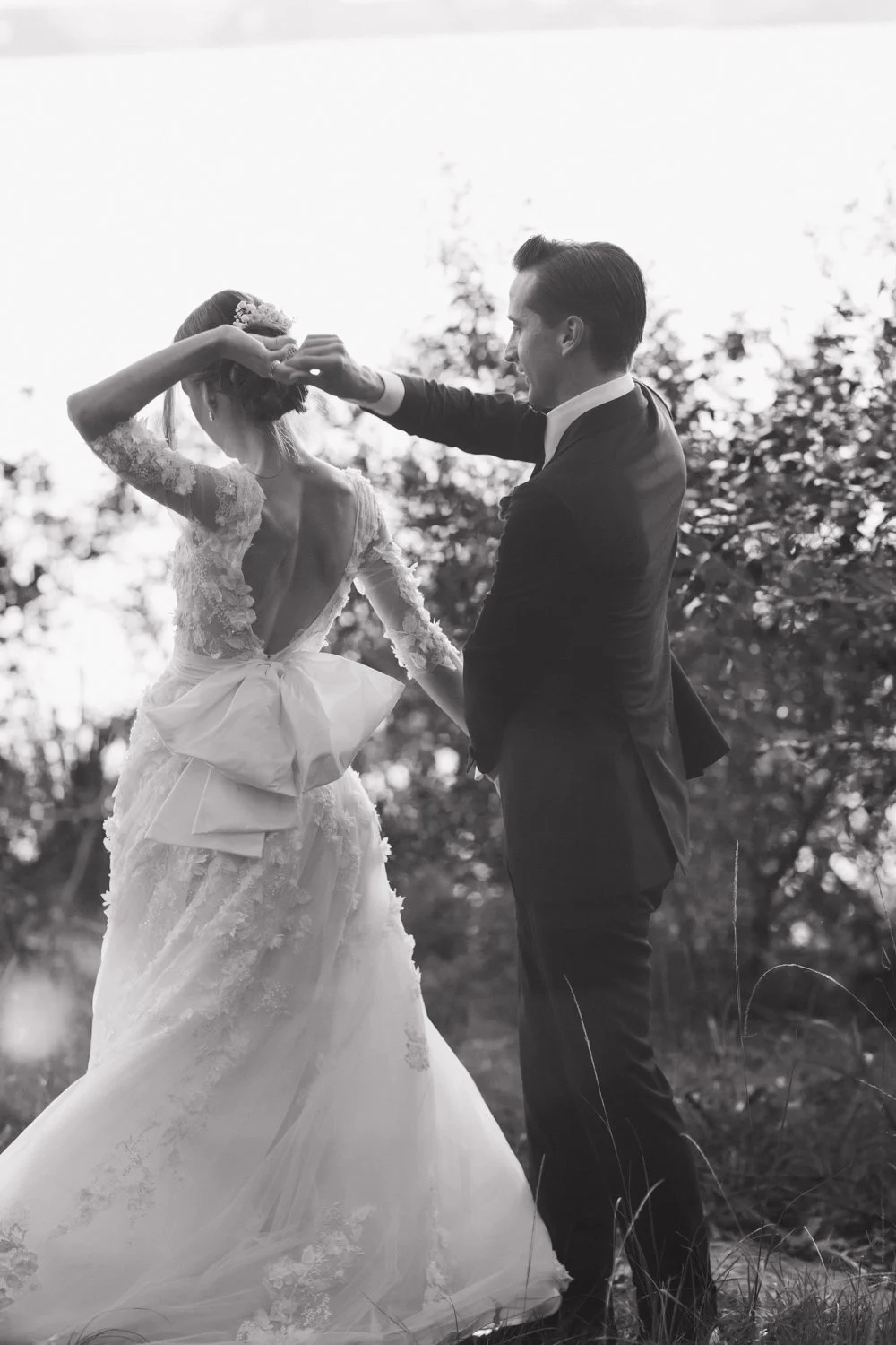 candid moment in black and white of bride and groom dancing in a grassy setting, bride turning her back showcasing her open-back gown with oversized bow.