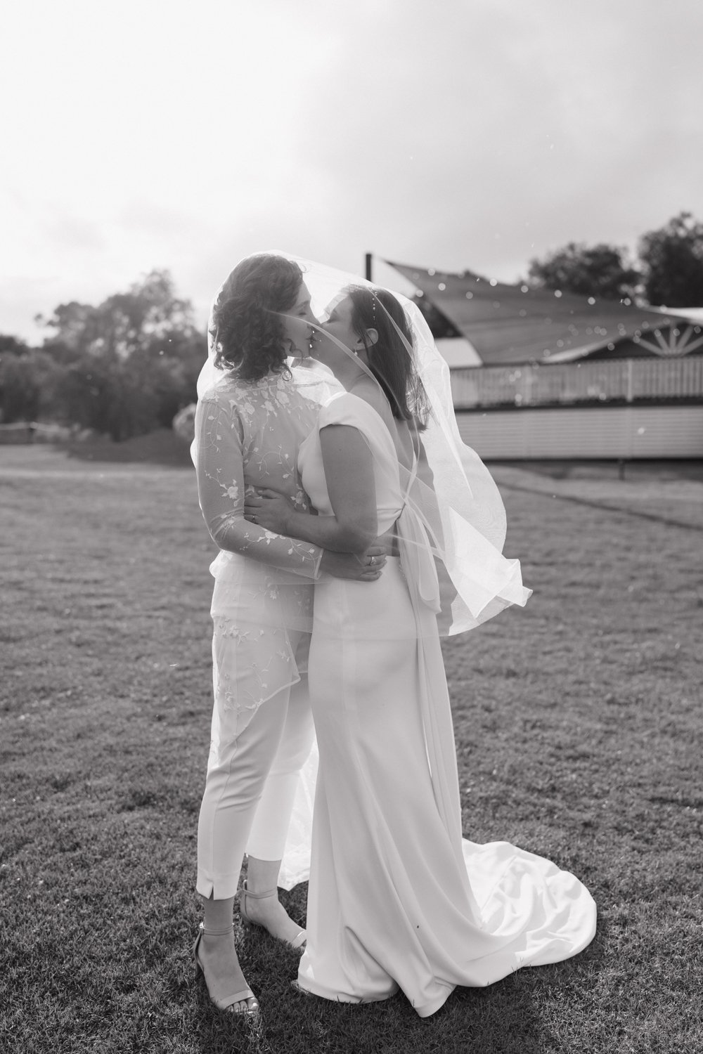 Two brides share a kiss under their veil that flows softly in the breeze, captured in timeless black and white