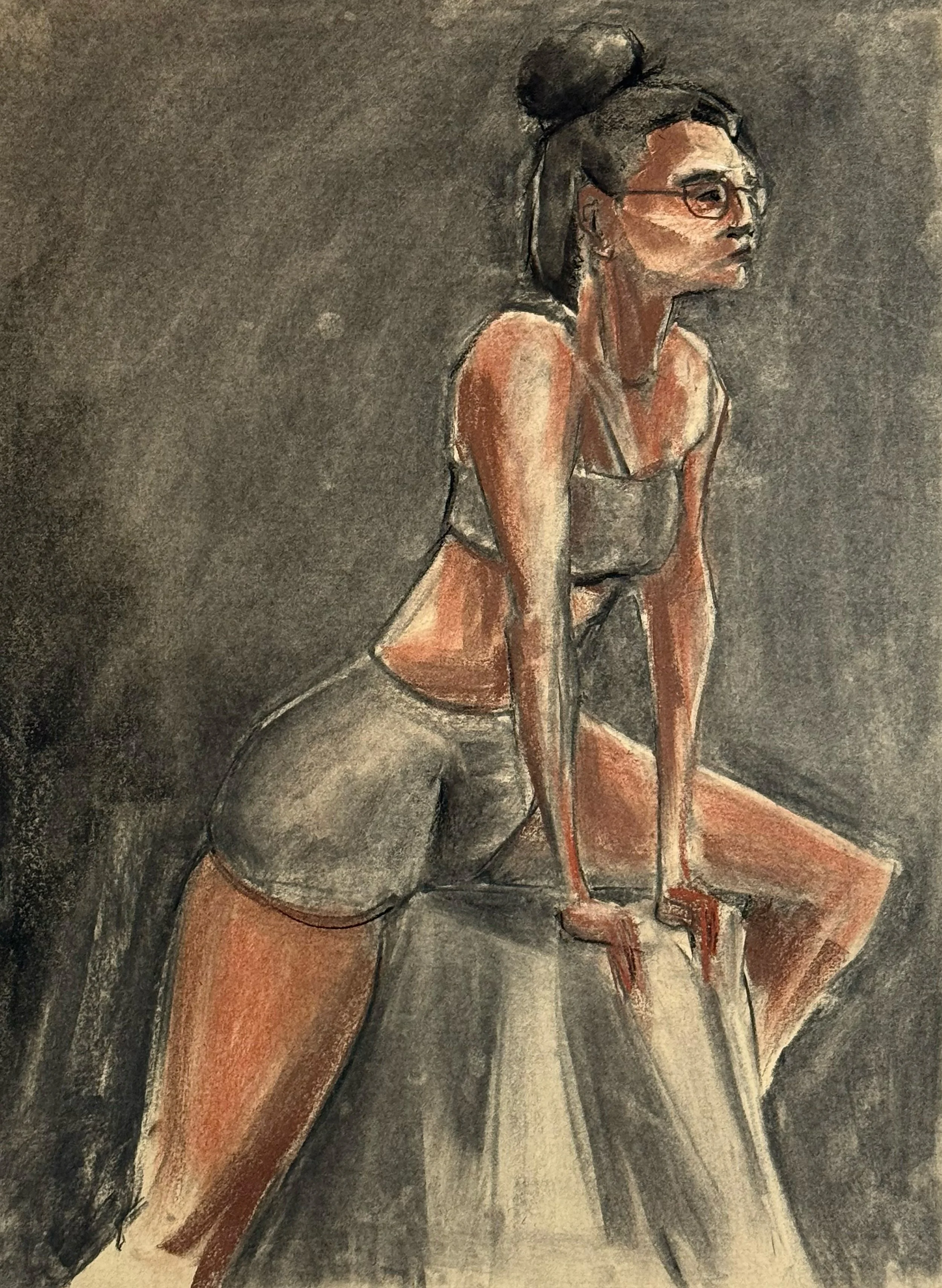A woman with glasses and her hair in a bun, leaning on a surface with her hands, wearing a sports bra and shorts, against a dark background.