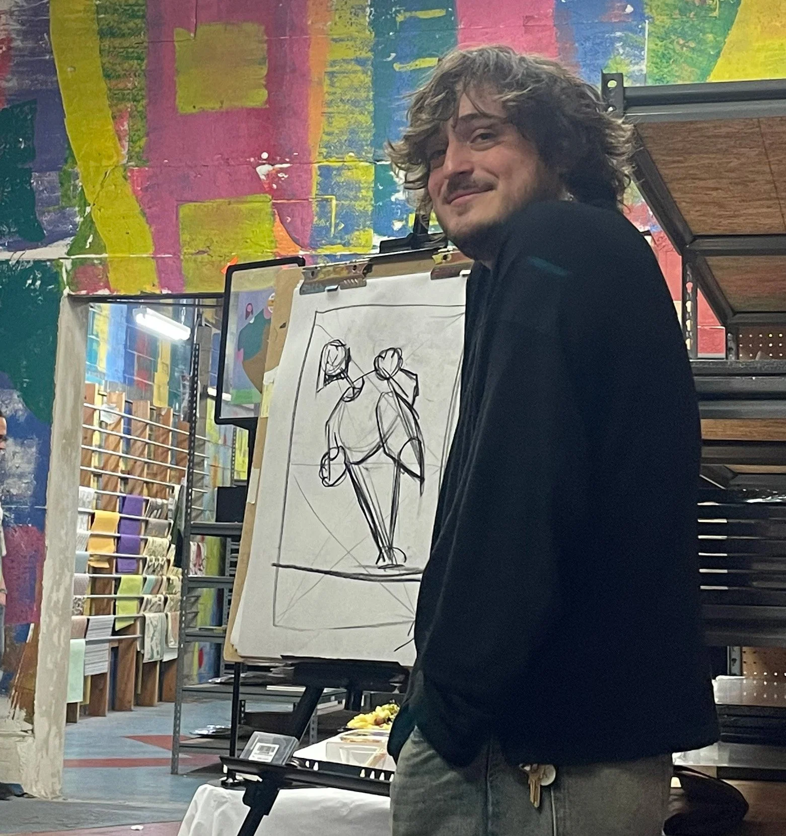 A man with curly hair and a beard standing in front of a sketch of two figures, inside an art store with colorful artwork on the walls and shelves of rolls of paper.