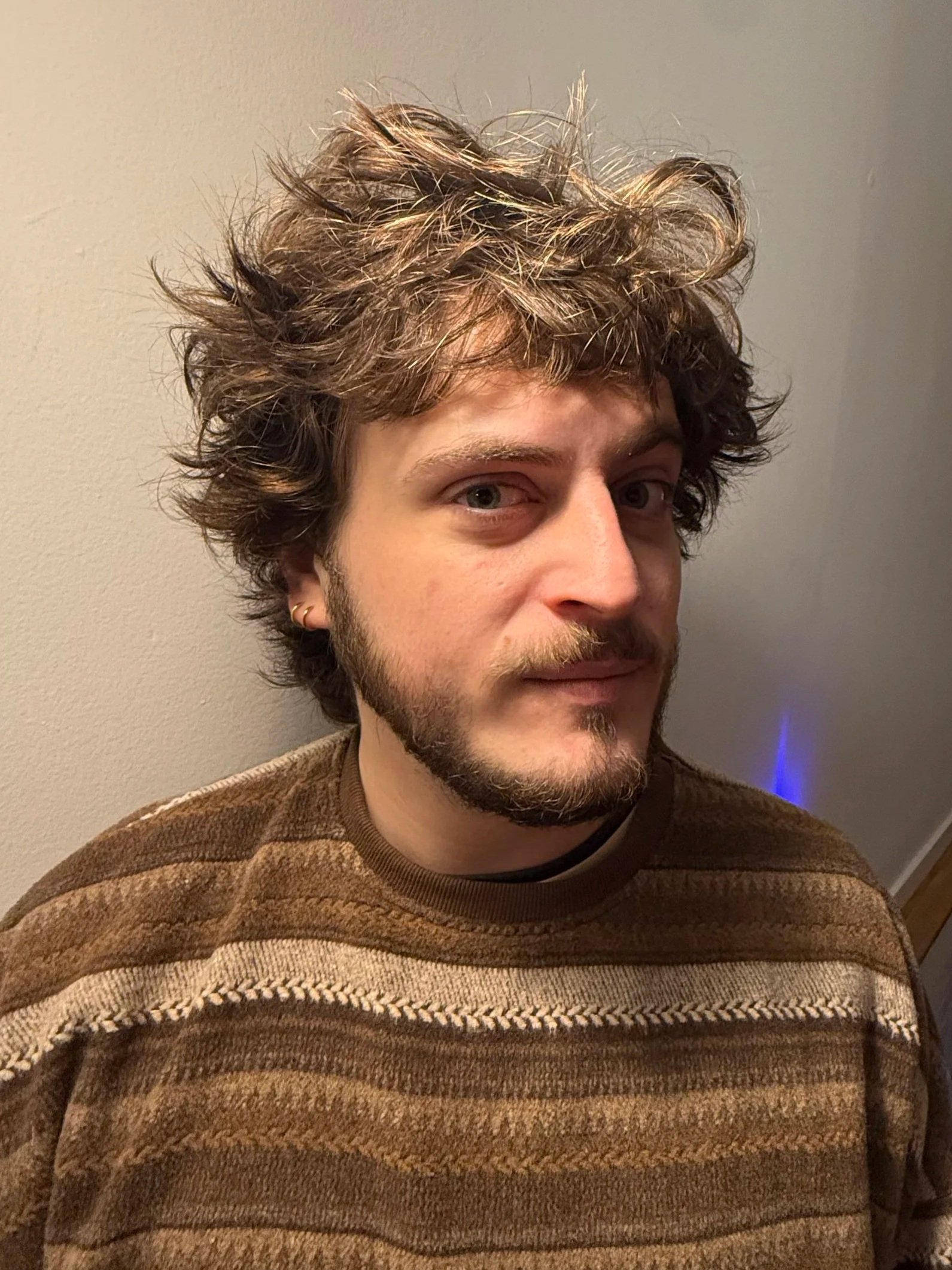 A young man with curly, disheveled hair and a beard, wearing a brown striped sweater, standing against a plain wall.