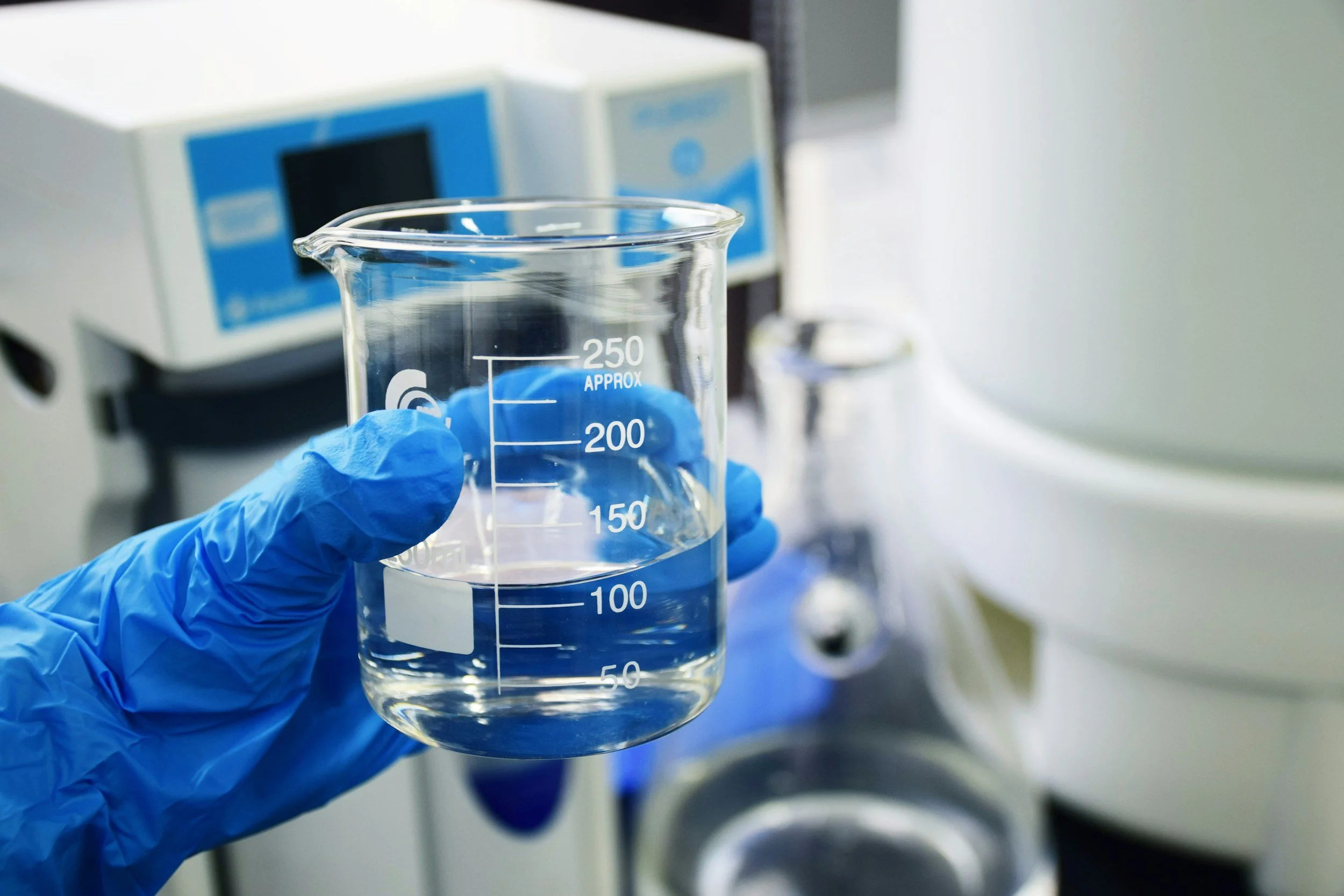 A scientist wearing blue gloves holding a glass beaker filled with a clear liquid in a laboratory.