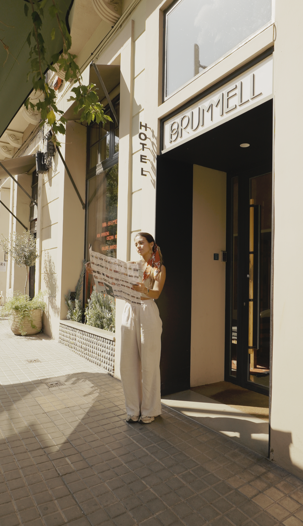 A woman stands outside the Brummell Hotel, reading a map on a sunny sidewalk with potted plants and large windows.