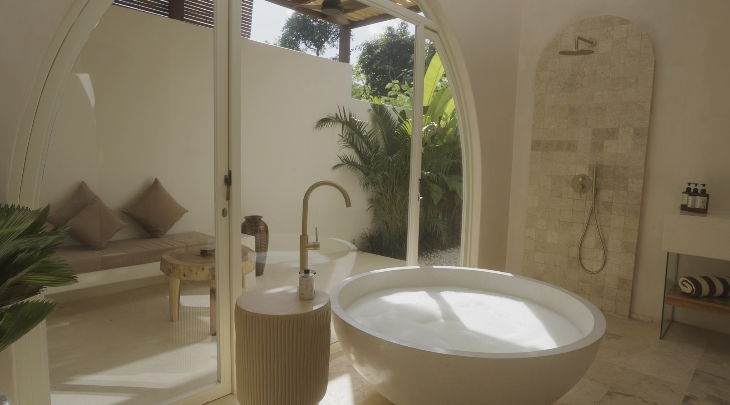 A luxurious bathroom with a freestanding bathtub, tiled shower, and a seating area outdoors with lush greenery visible through large glass doors.