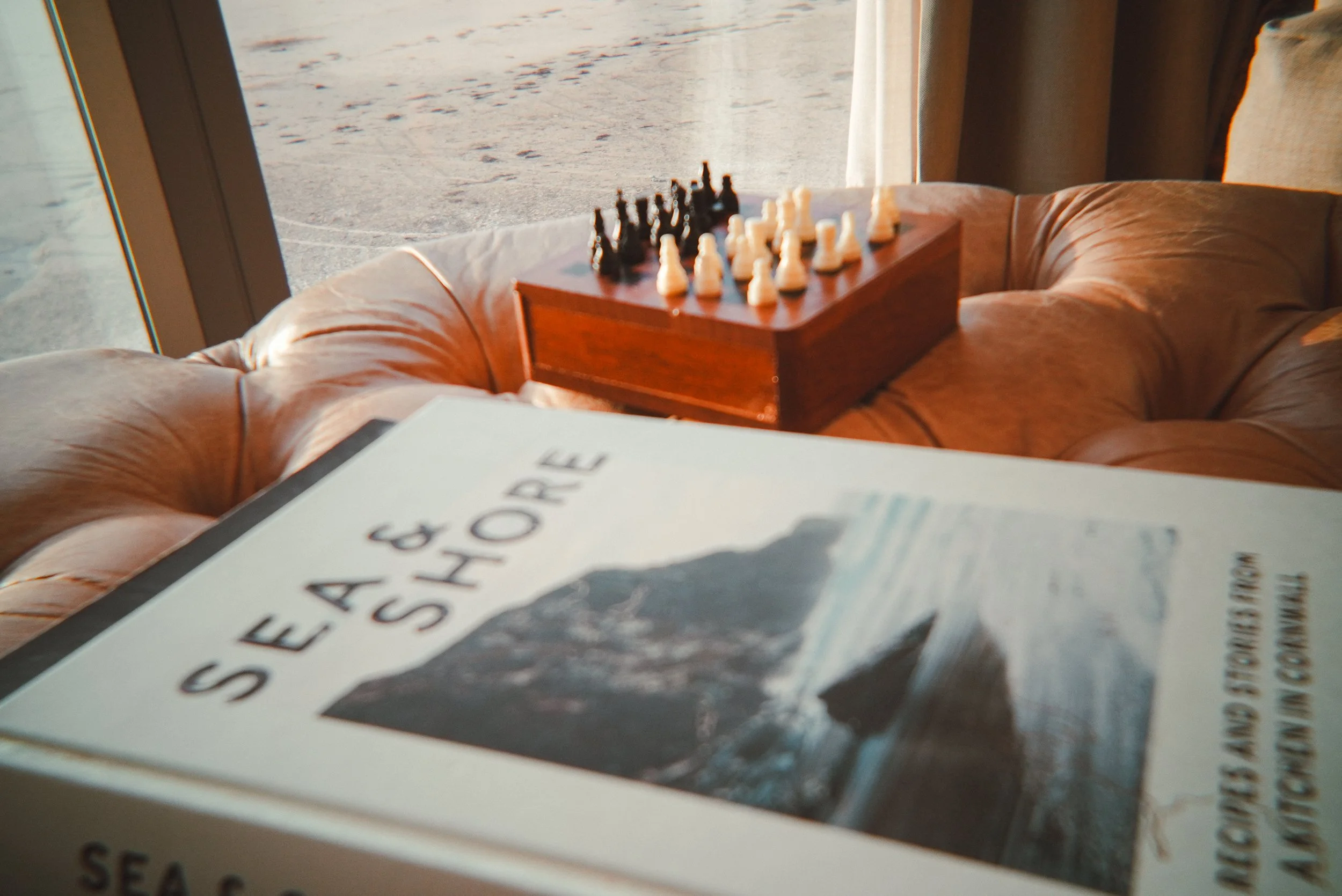 A chessboard set up, along with a book titled 'Sea & Shore' on a leather sofa near a window revealing an outdoor landscape.