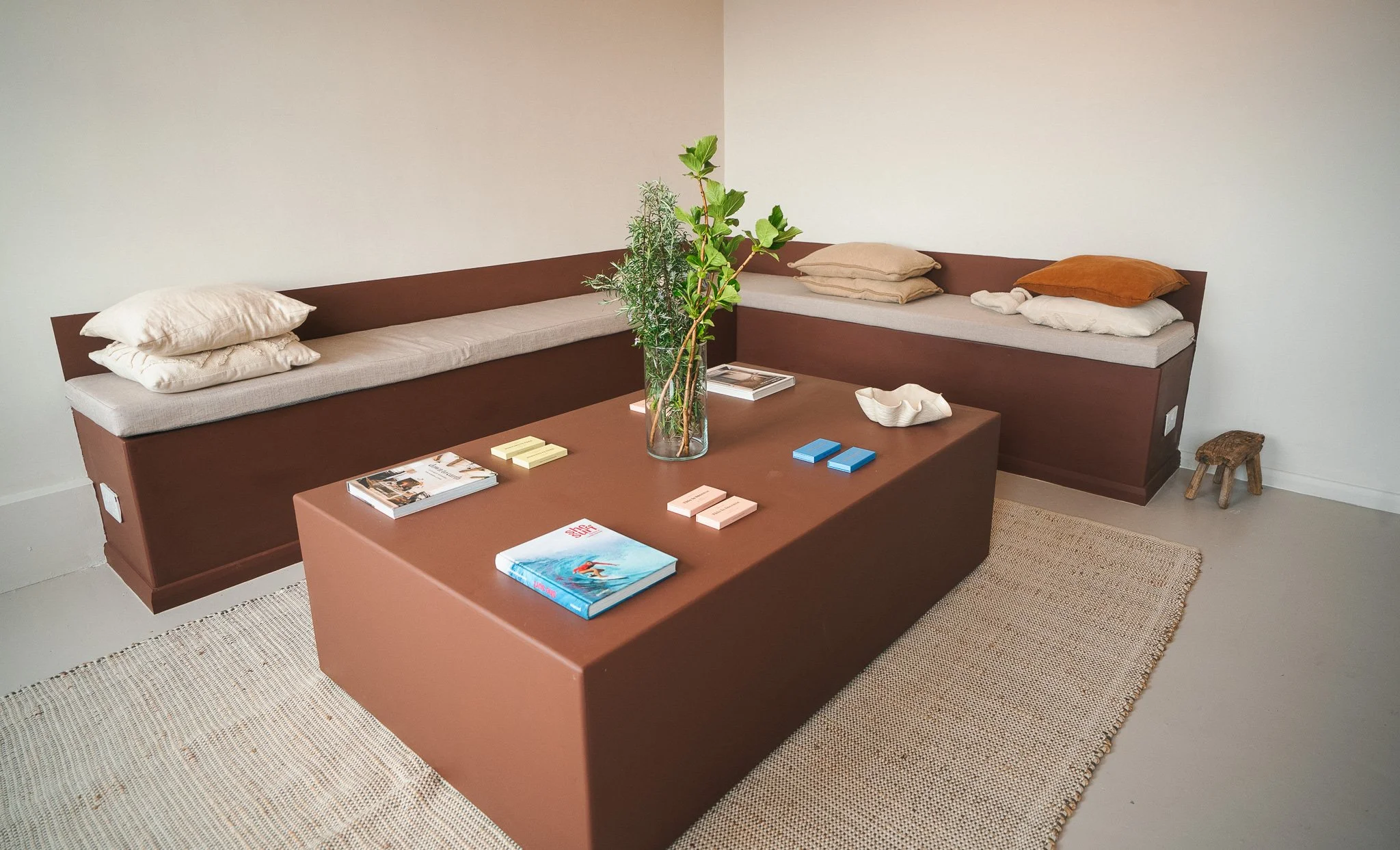 Pilates studio reception area with a brown sofa,  a central brown coffee table with books, magazines, and a decorative bowl, and a potted plant in the middle. The room has light-colored walls, a beige rug, and a small wooden stool in the corner.