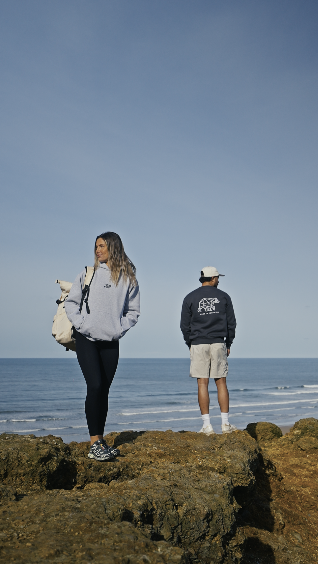 A woman and a man standing on rocks by the beach, looking at the ocean, with a clear blue sky overhead modelling for a clothing brand.