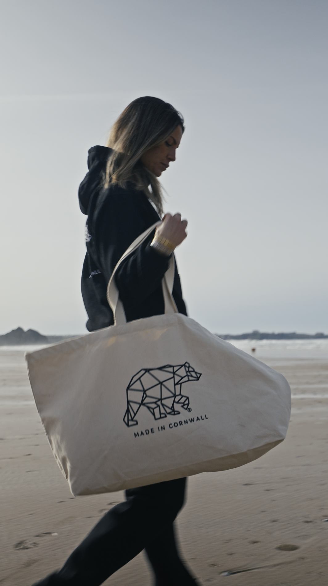 A woman walking on the beach carrying a tote bag with a geometric bear logo and the text 'Made in Cornwall' modelling for a clothing brand.