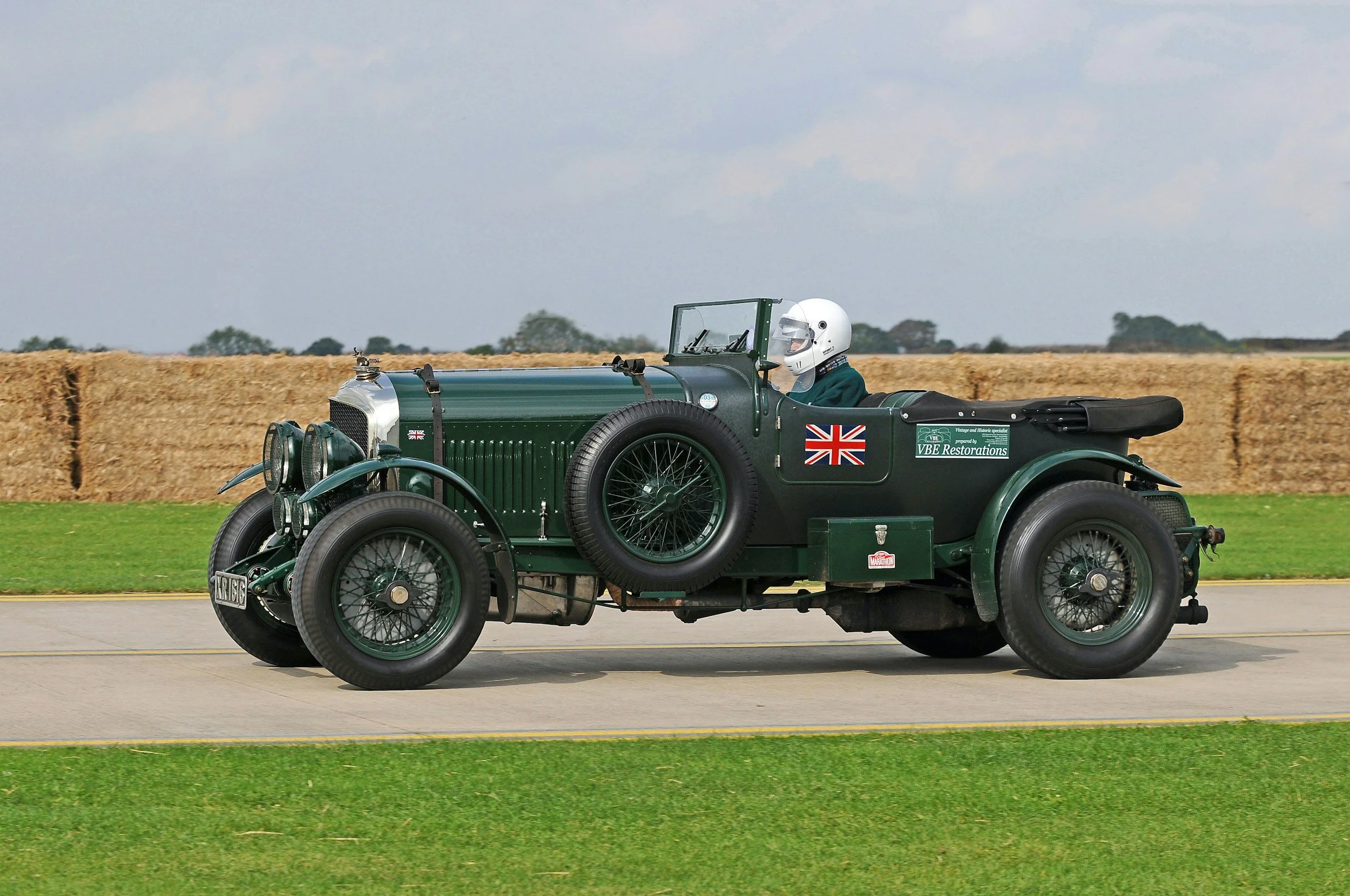 Vintage green open-top racing car with British flag decals, driven by a person wearing a white helmet, on a track with a hay bale barrier and grassy field in the background.
