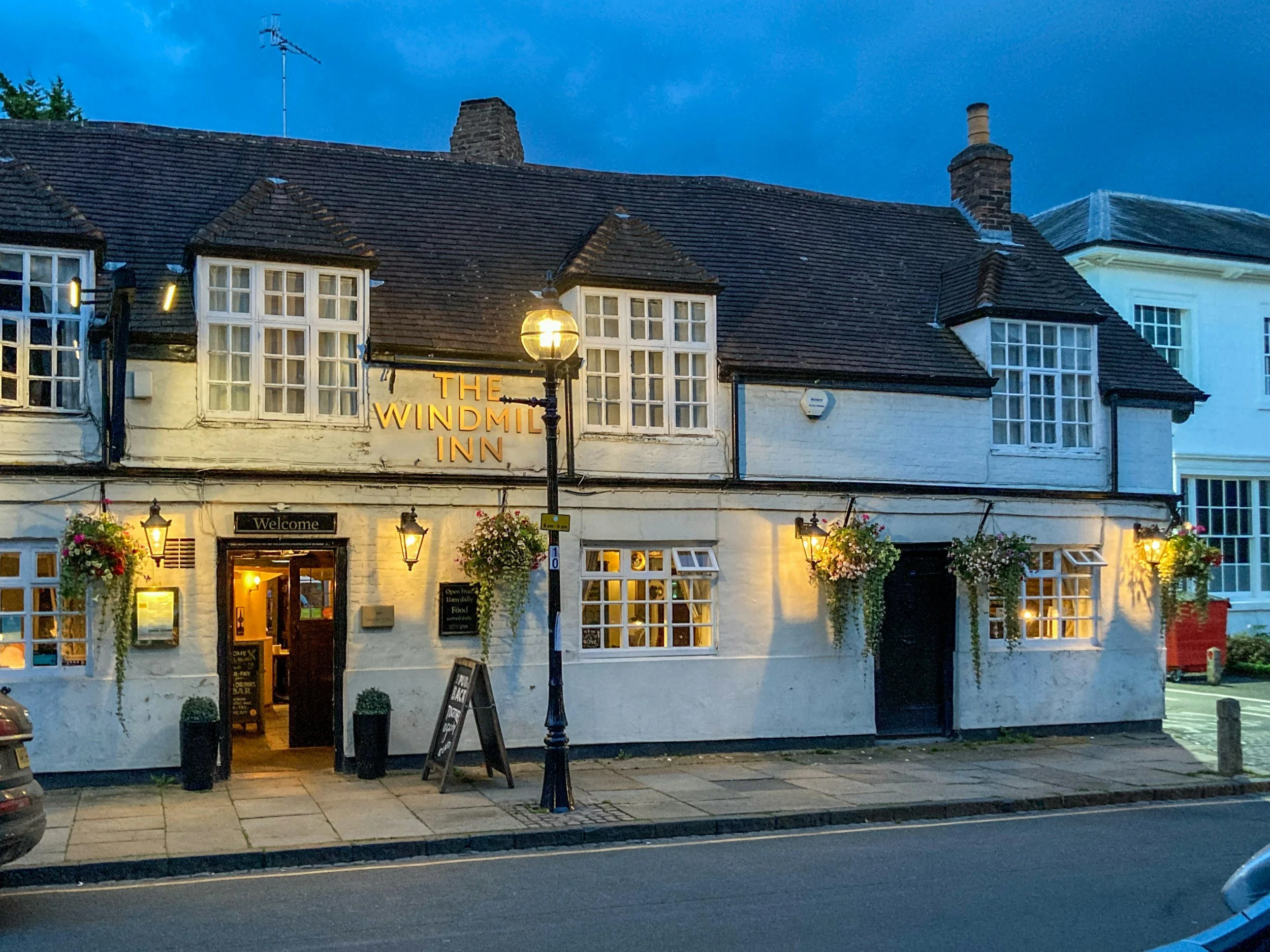Exterior view of The Windmill Inn, a cozy pub with hanging flower baskets, warm lighting, and a welcoming entrance on a calm evening.