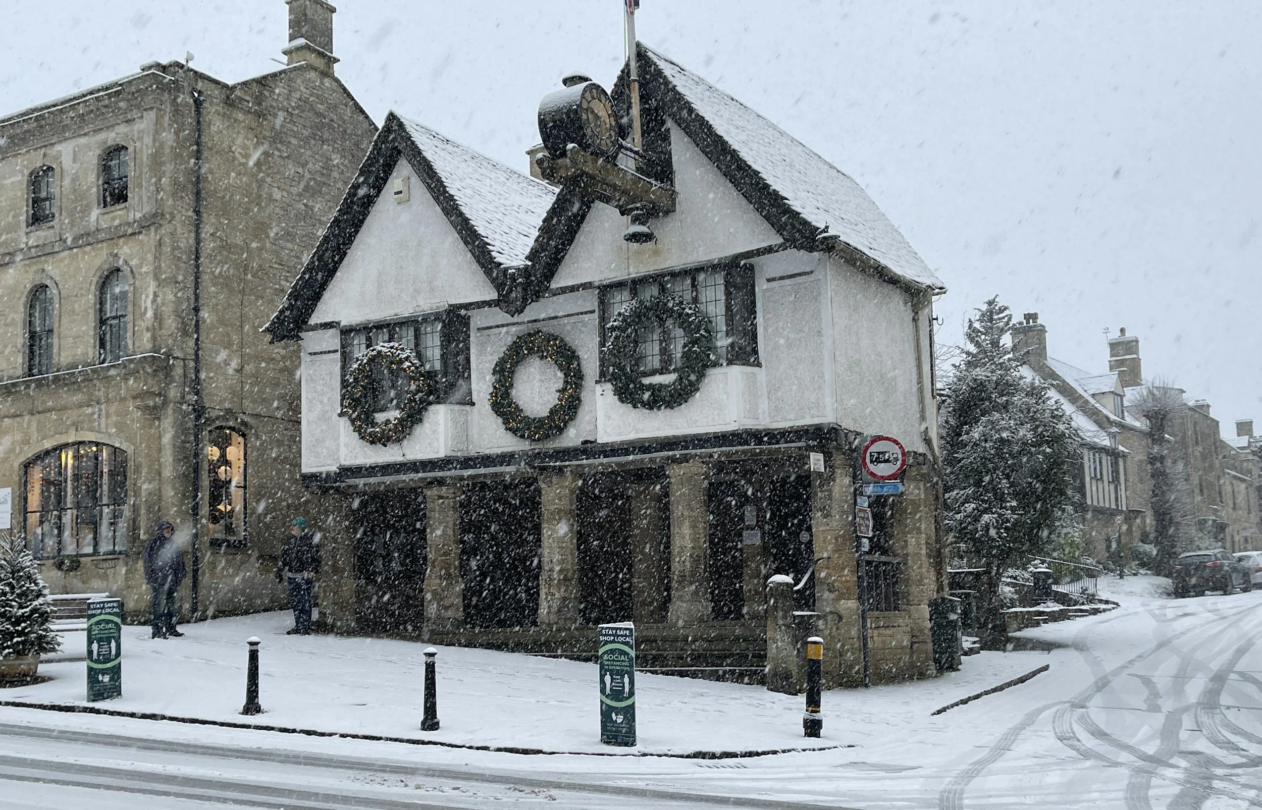 A snowy street scene with a white building decorated with holiday wreaths and a large clock on the roof, snow falling heavily, and a few pedestrians walking.