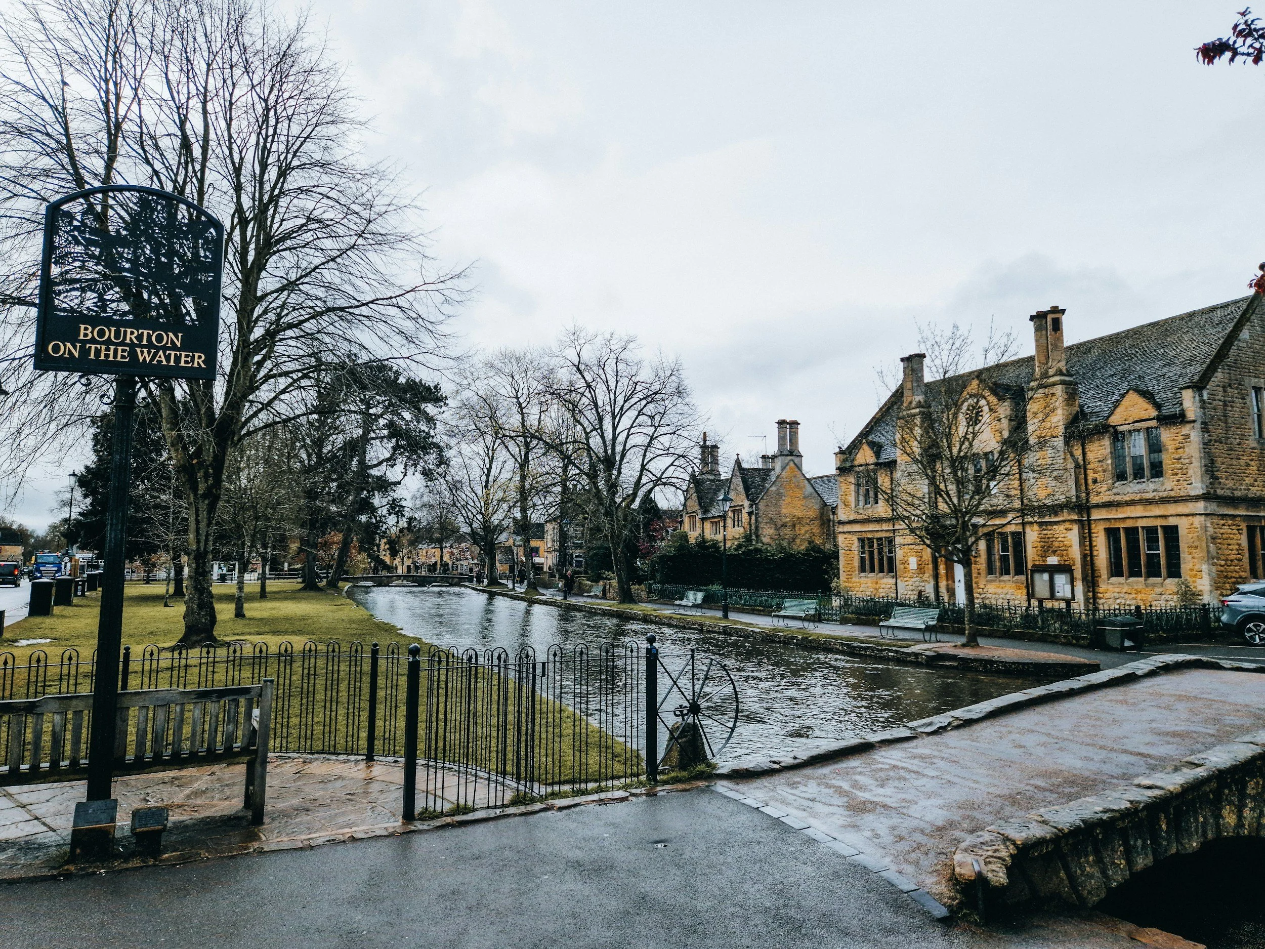 Scenic view of a small canal with historic stone buildings on the right and a park with trees on the left under an overcast sky, in Bourton on the Water, England.