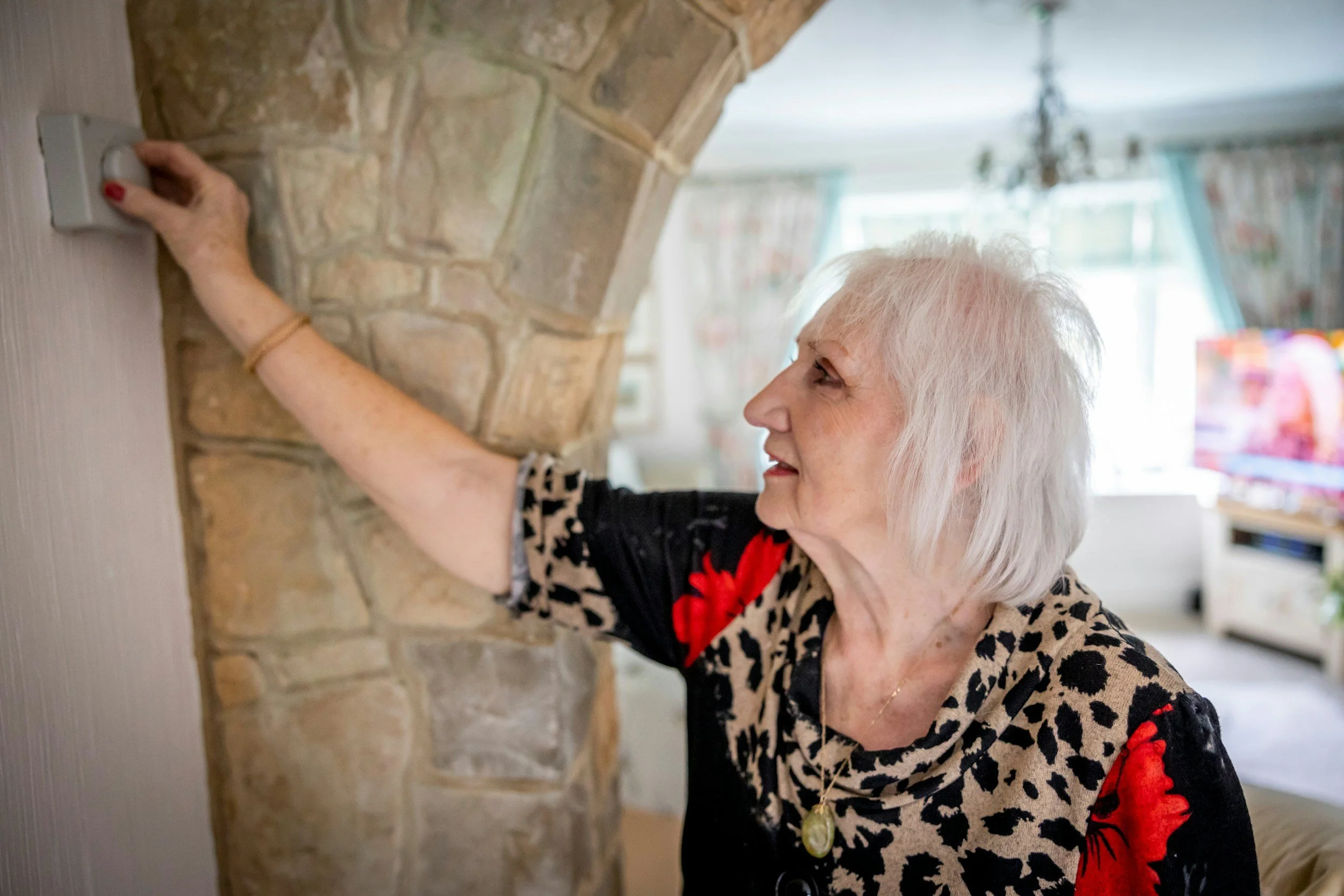 An elderly woman with white hair adjusting a wall thermostat in a living room with a stone fireplace, floral curtains, and colorful decorations in the background.