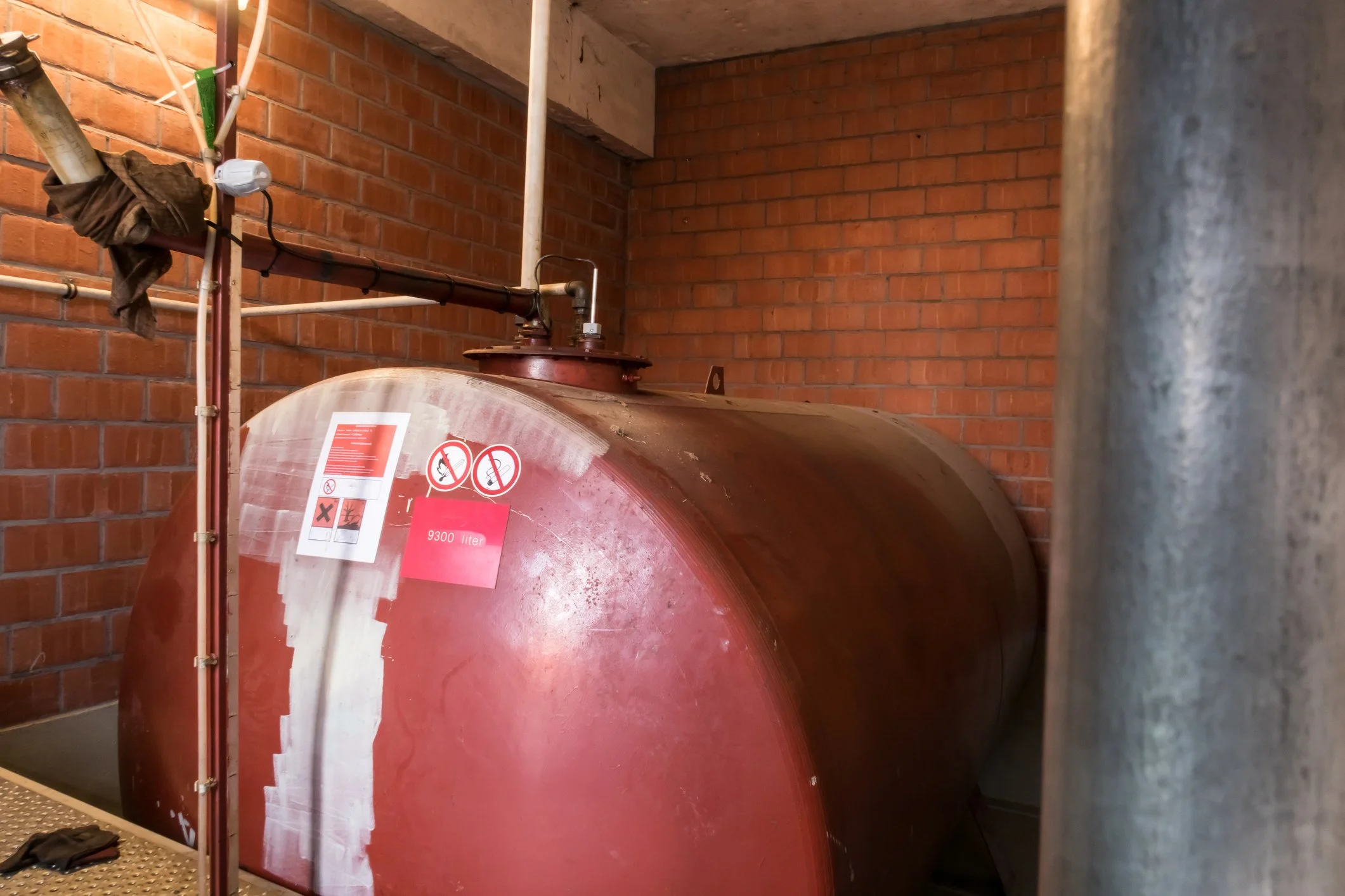 Large brown metal tank in a room with brick walls, with warning labels and pipes attached.