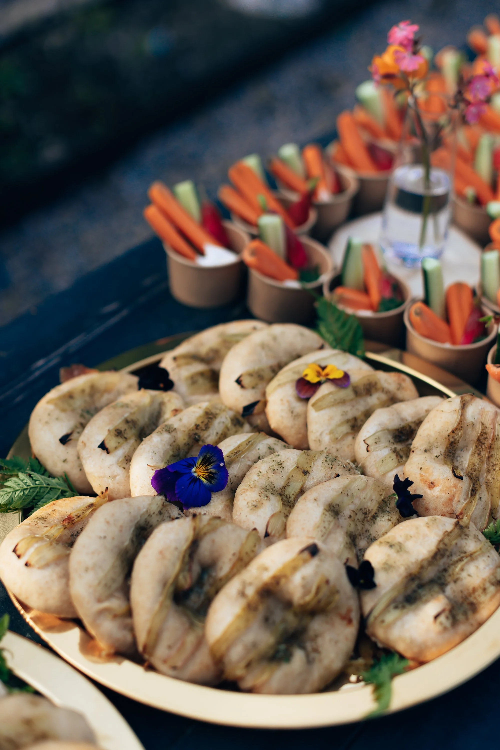 A platter of small sandwiches garnished with edible flowers, with a background of small cups filled with vegetables and dip, arranged for a catering event.