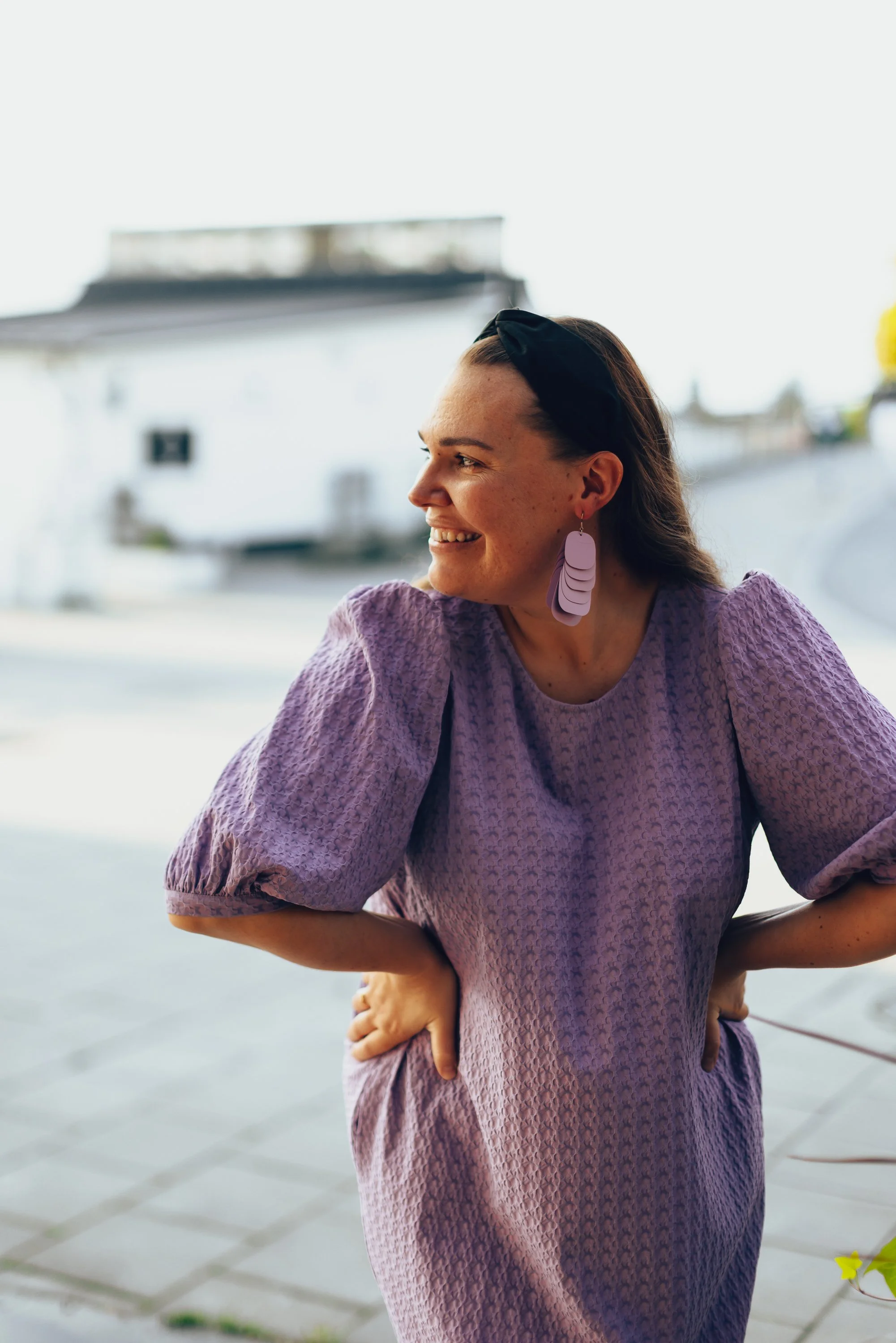 A woman in a textured purple dress with puffed sleeves, smiling and looking to the side, wearing large purple earrings and a black headband, outdoors on a sunny day.