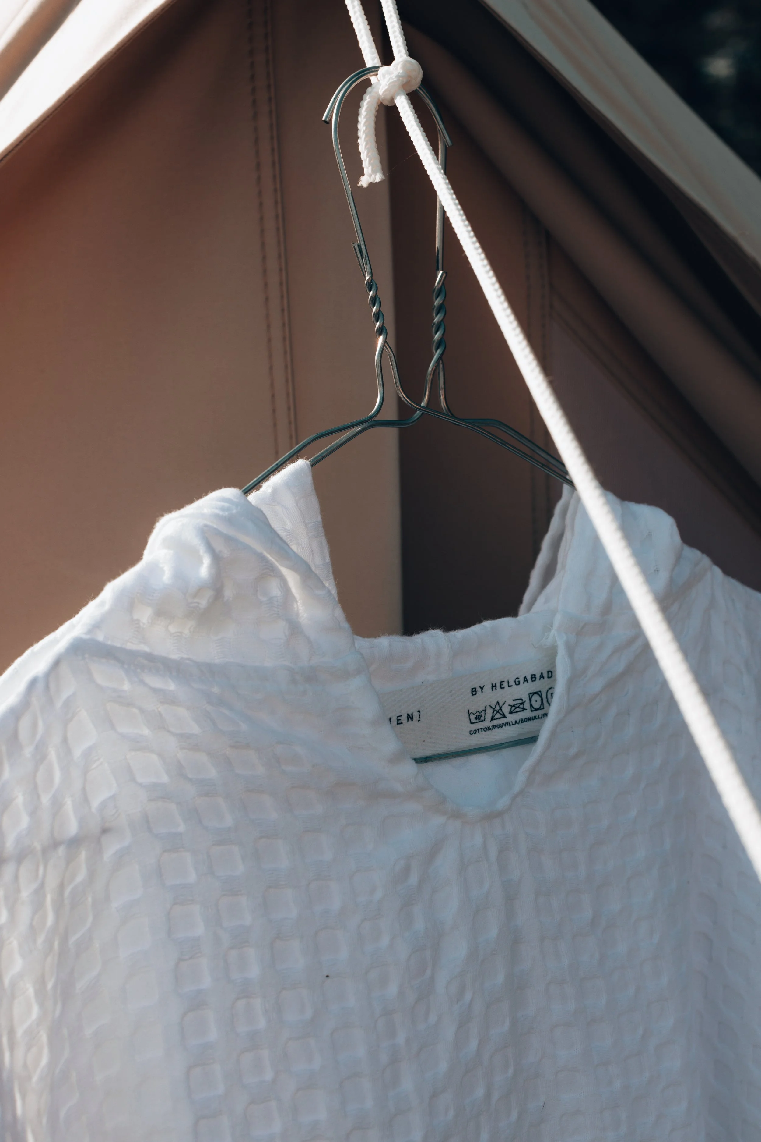 A white textured blouse hanging on a metal hanger, attached to a white rope, inside a closet.
