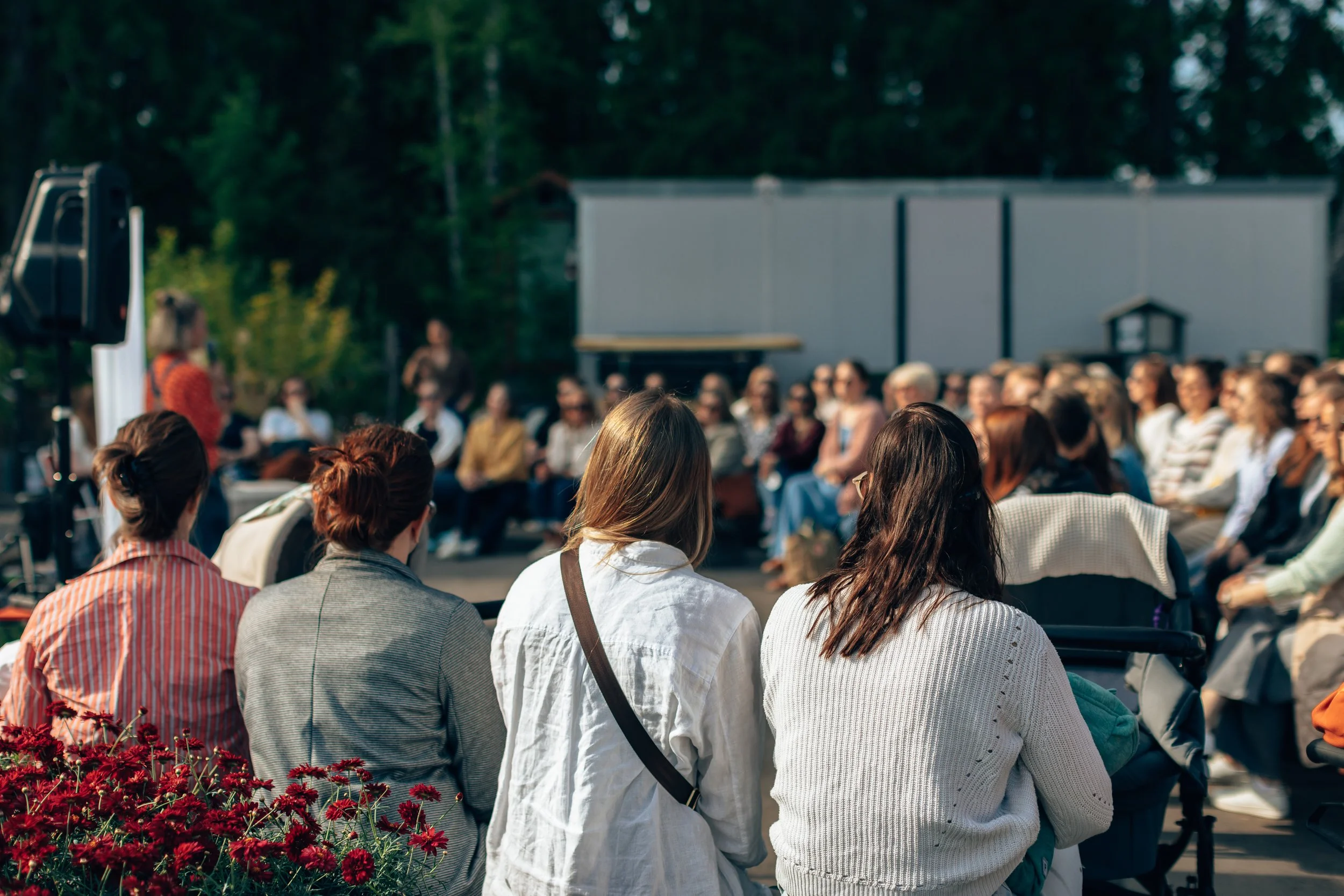 A large outdoor gathering of people sitting in a circle, engaged in an event or discussion, with trees in the background and a building or shed behind them.