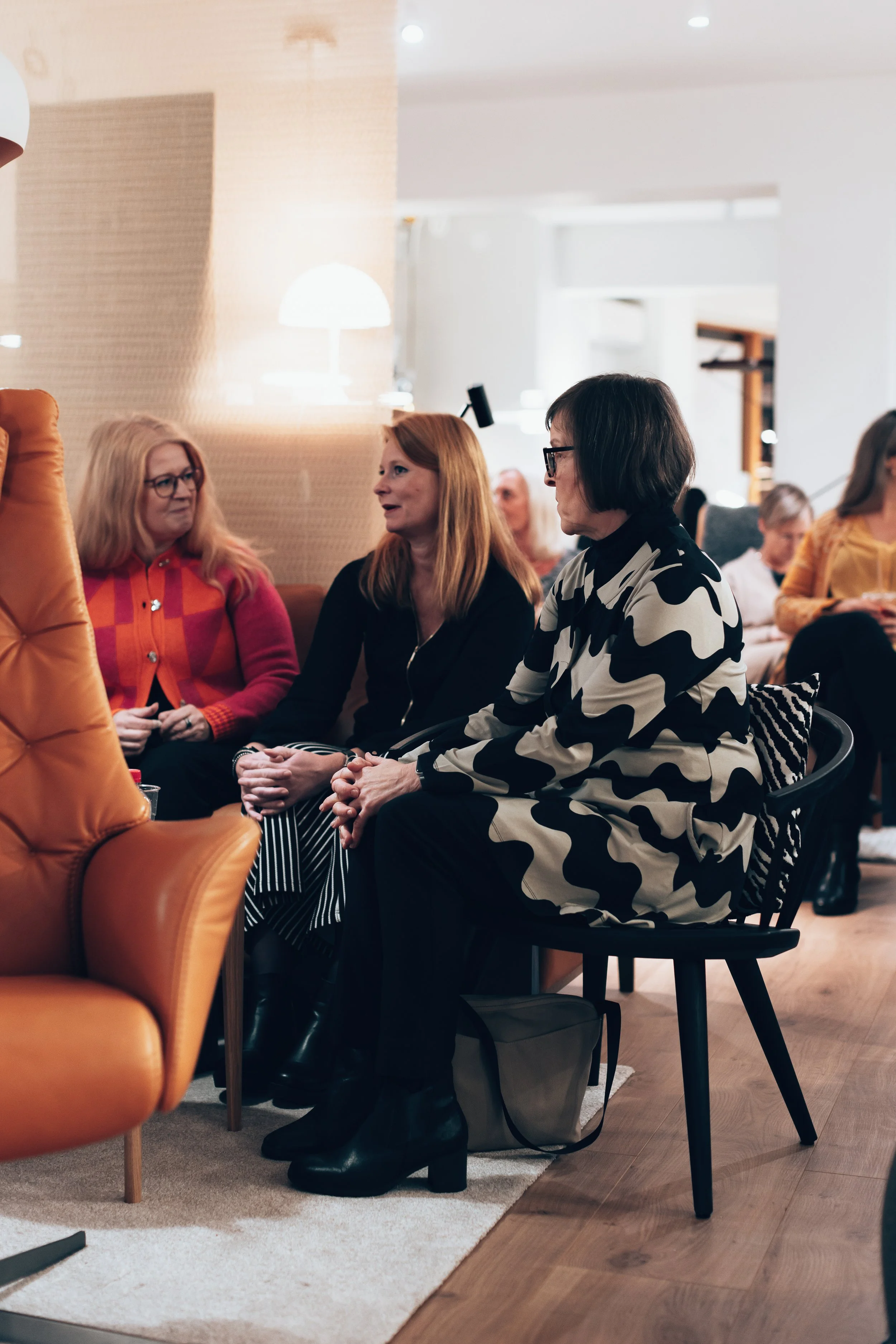 Three women sitting and talking in a cozy indoor setting, with other people in the background, featuring warm lighting and modern decor.