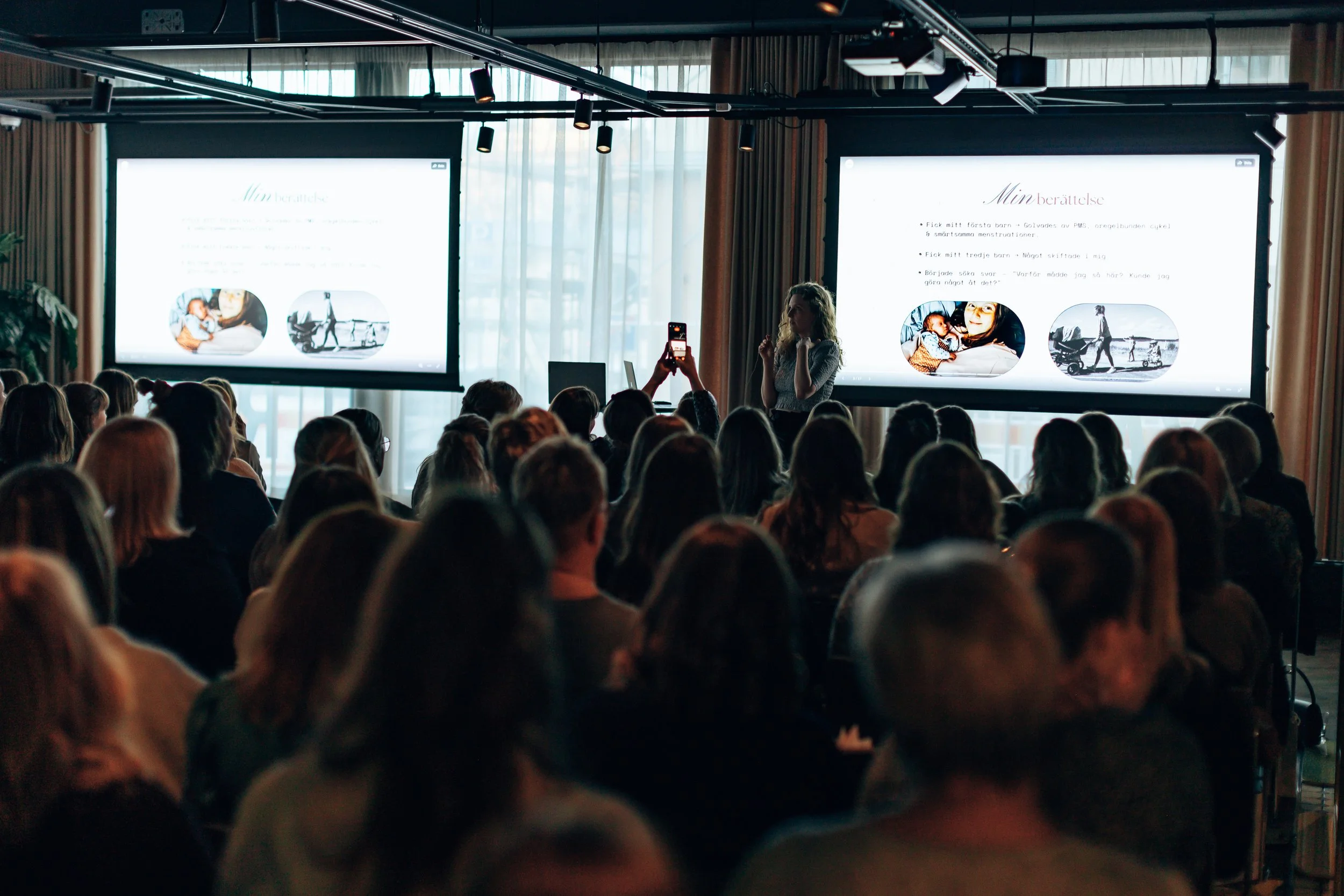 A woman giving a presentation to a large audience seated in a dimly lit room with two large projection screens displaying slides, and a person in the audience taking a photo with a smartphone.