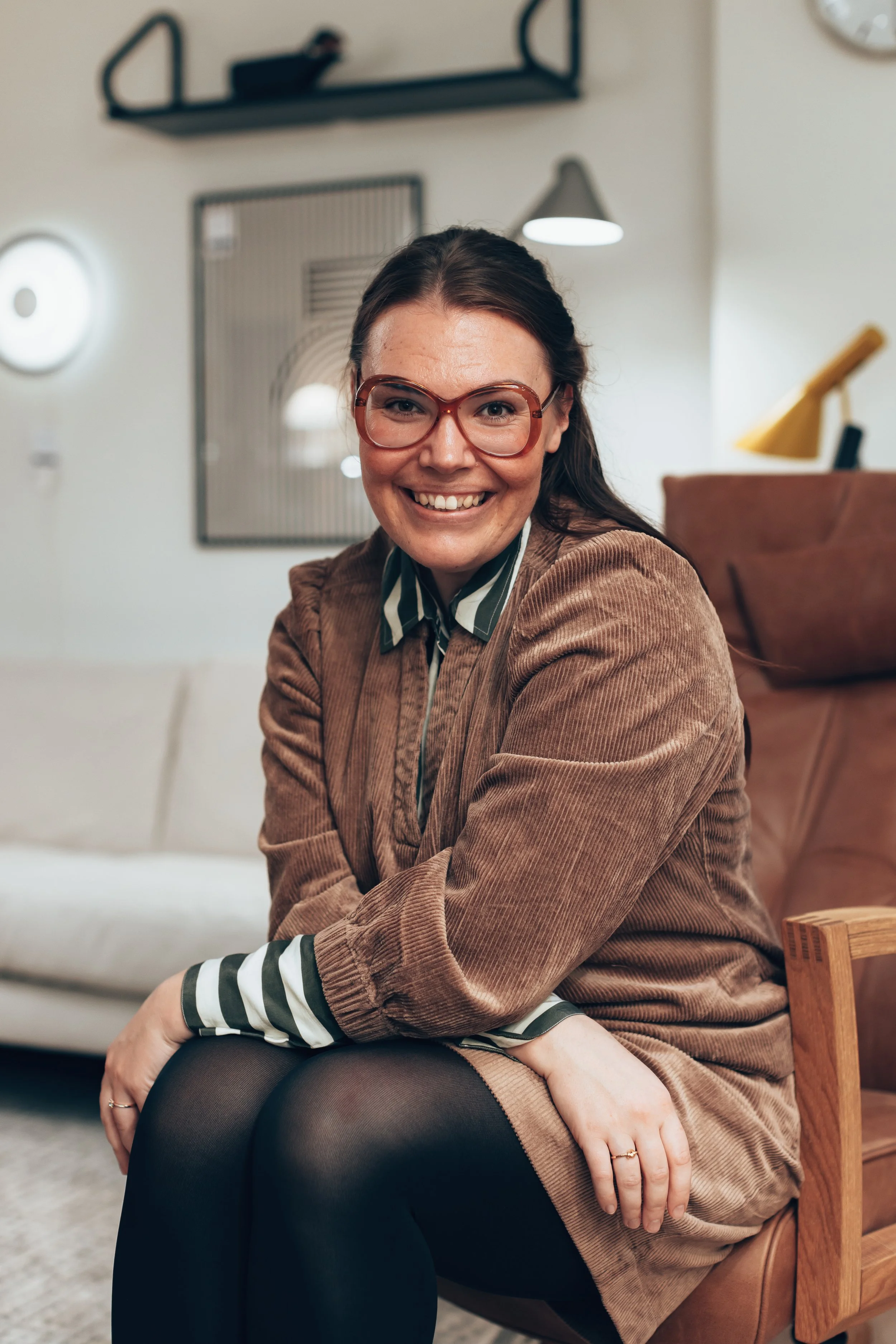 A woman with glasses smiling, sitting on a wooden chair in a cozy, modern living room.