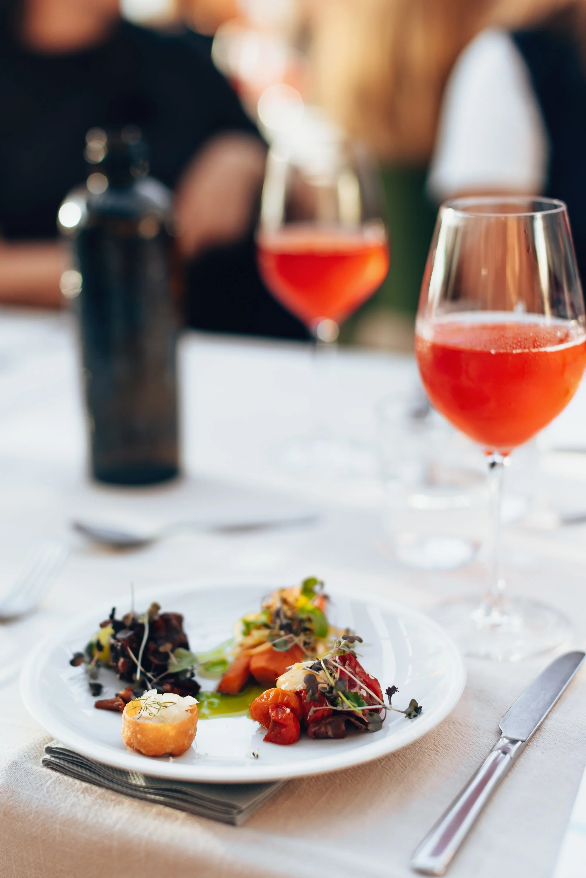 Appetizer plate with various small dishes, two glasses of pink cocktail, and a dark bottle on a white tablecloth.