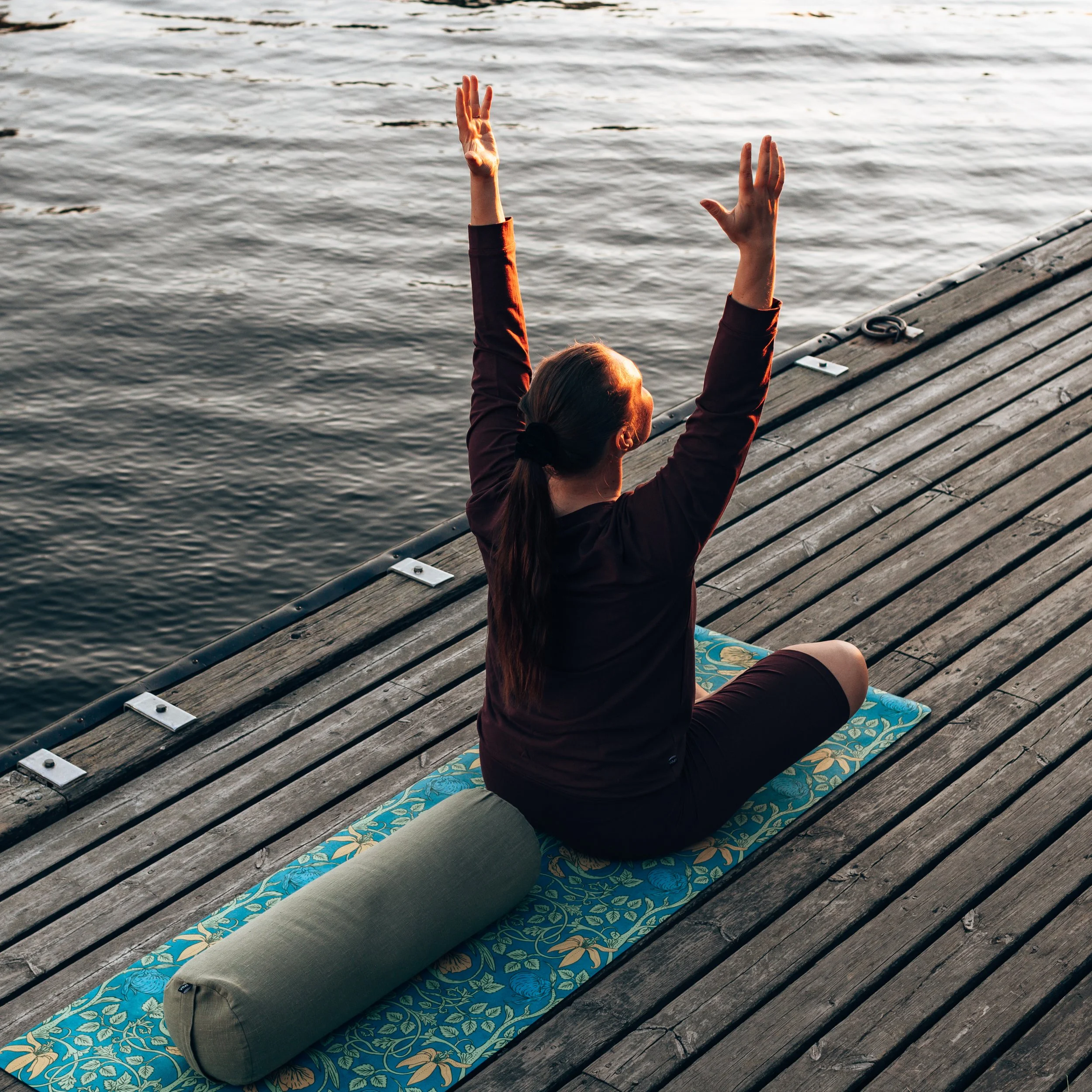 A woman practicing yoga on a mat on a wooden dock by the water, sitting cross-legged with arms raised.