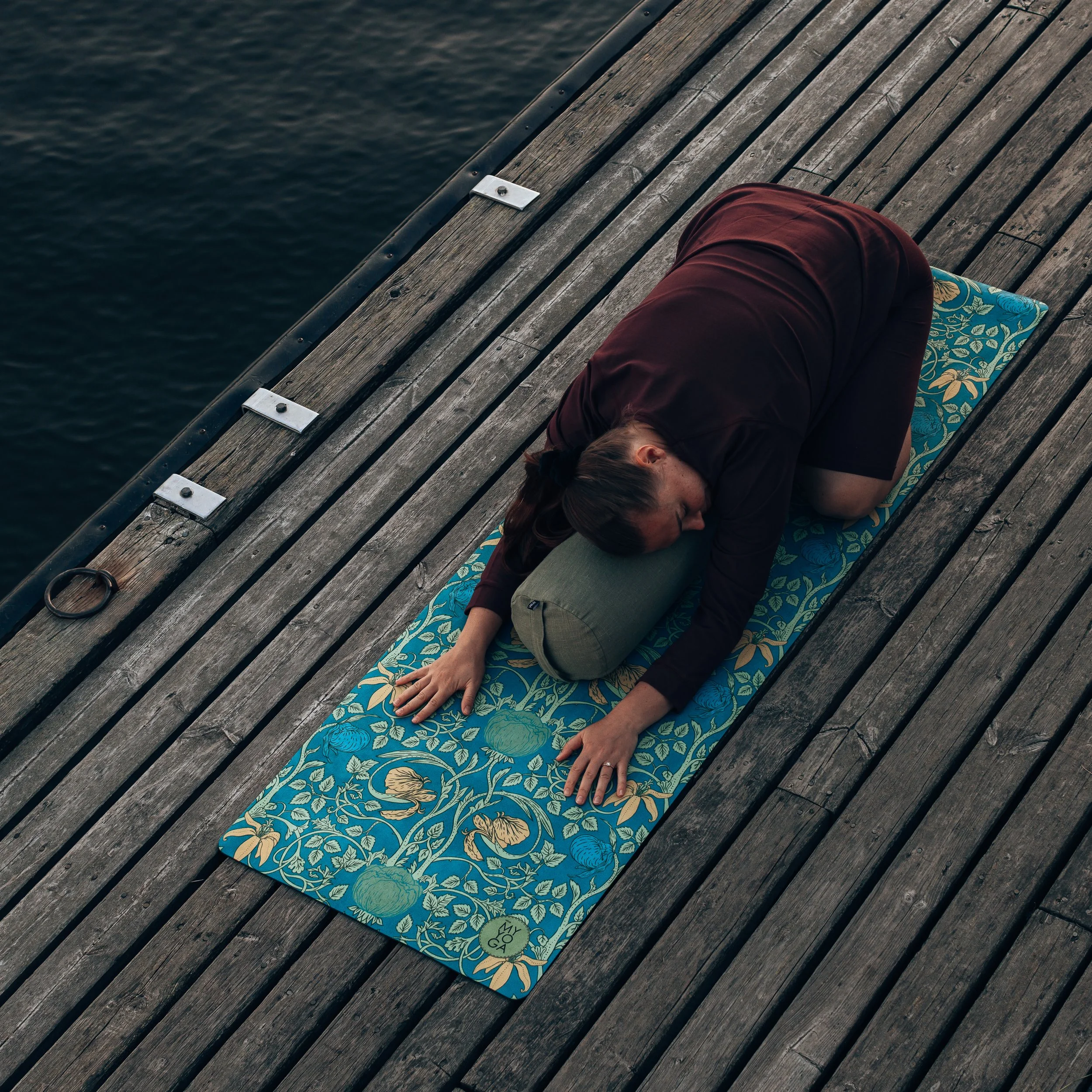 A woman practicing yoga on a patterned mat on a wooden dock next to water, using a bolster for support in a child's pose.