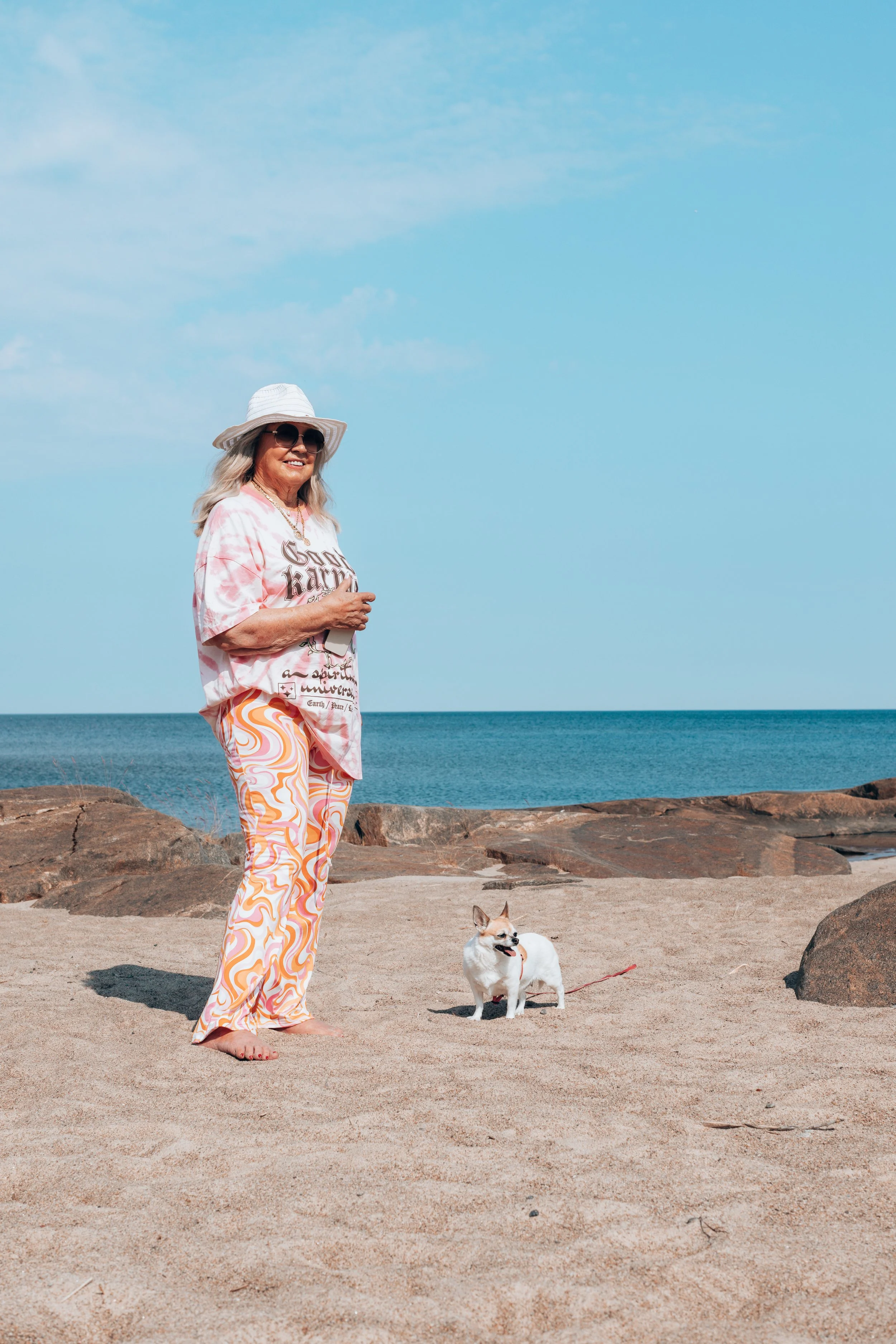A woman in summer clothing, including a white floppy hat, sunglasses, a pink and white tie-dye t-shirt, and colorful patterned pants, stands on a sandy beach with her small dog, near rocks with the ocean and sky in the background.