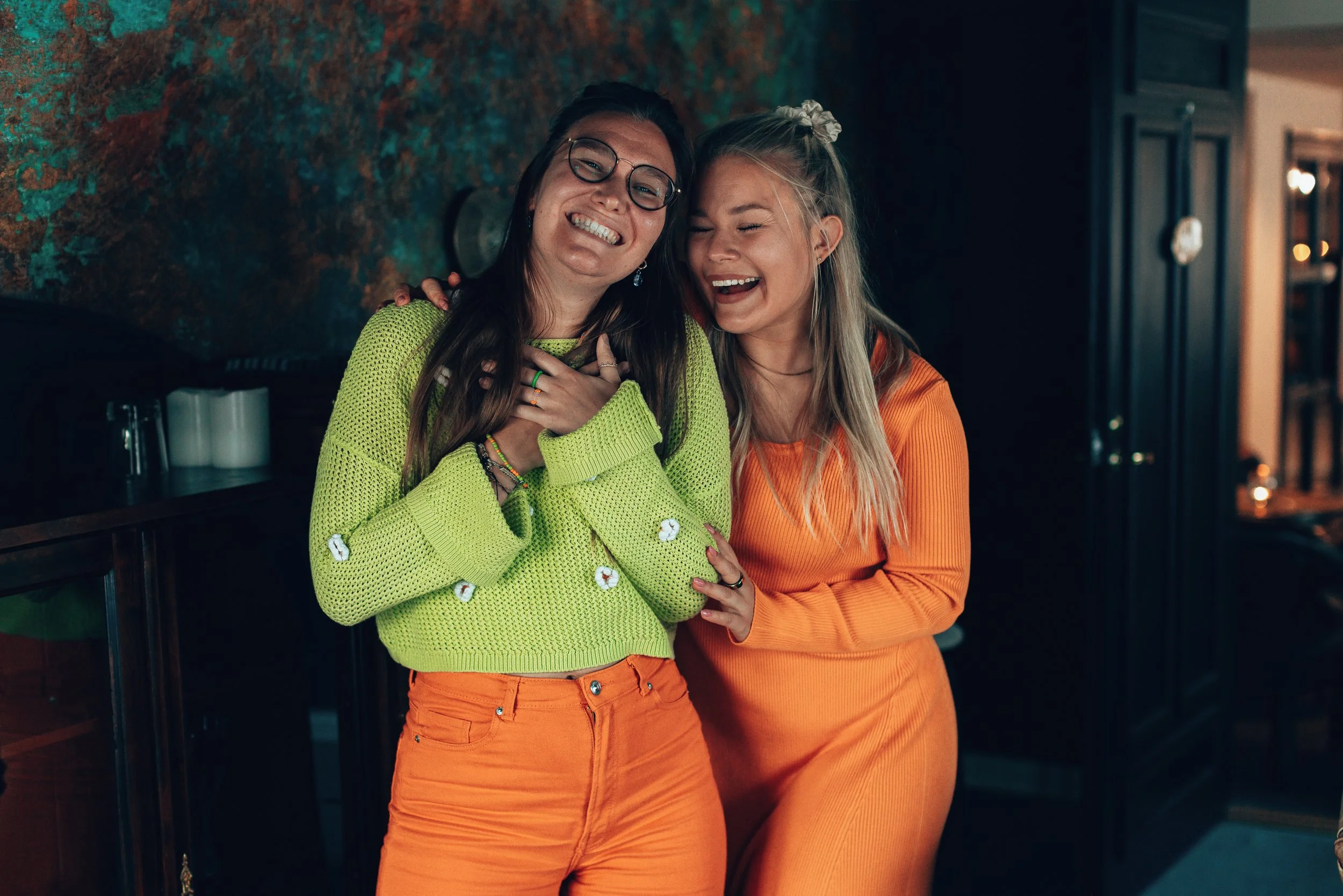 Two women smiling and laughing together indoors, one wearing a bright green sweater and orange pants, the other wearing an orange top and pants. They seem to be enjoying a happy moment.