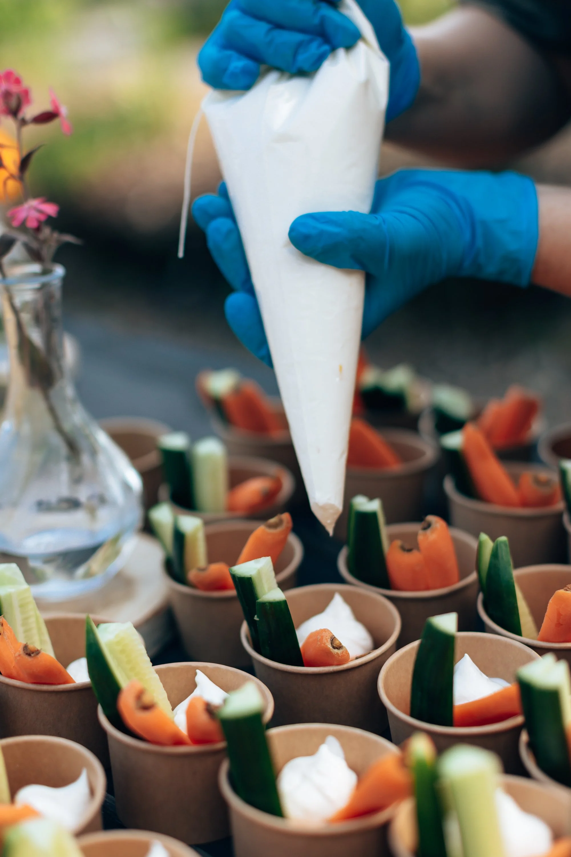Person wearing blue gloves piping a white creamy mixture into small cups filled with sliced carrots, cucumbers, and celery, with a blurred background.