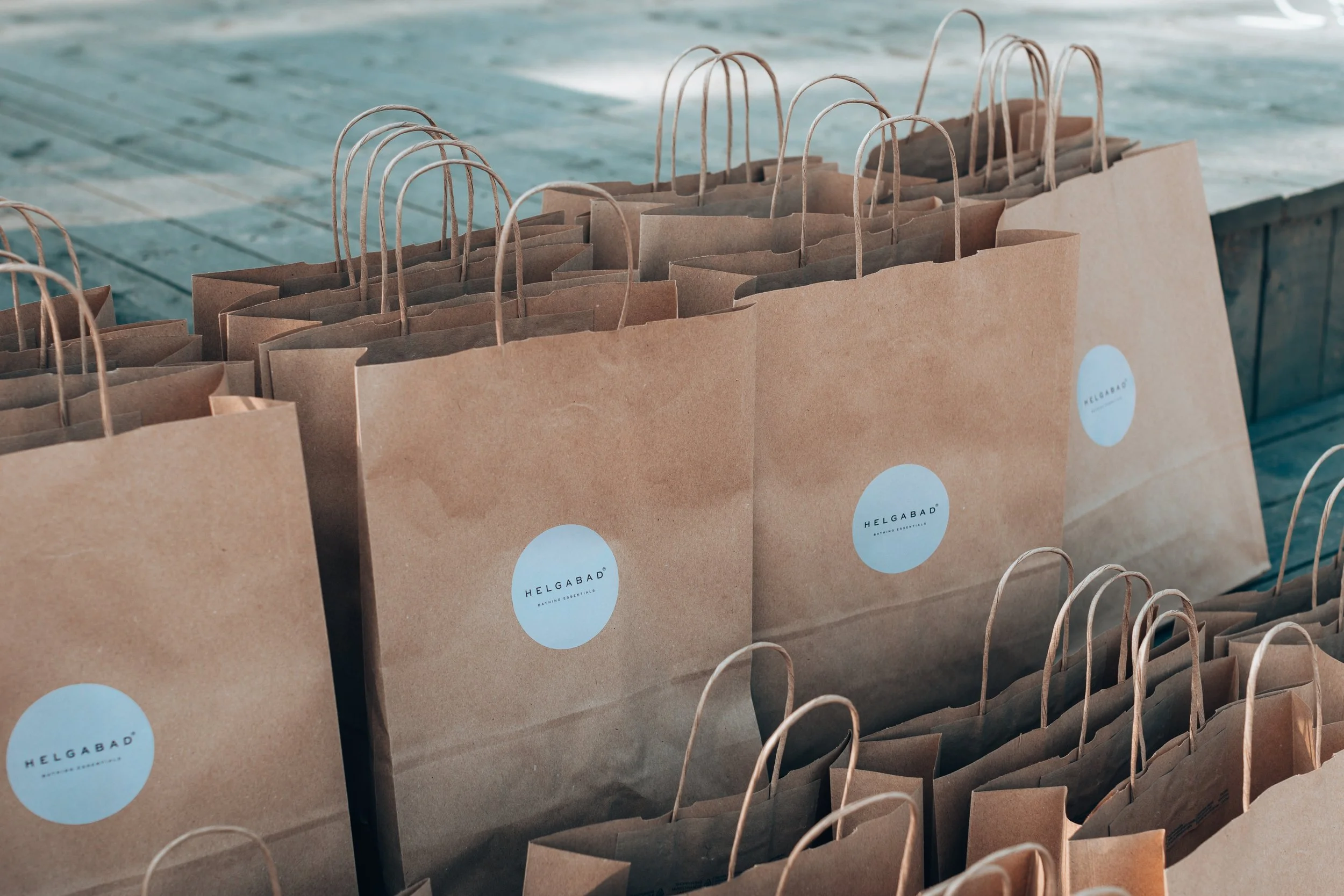 Multiple brown paper shopping bags with rounded handles and a white circular logo that reads 'HELGBAD' on a wooden surface.