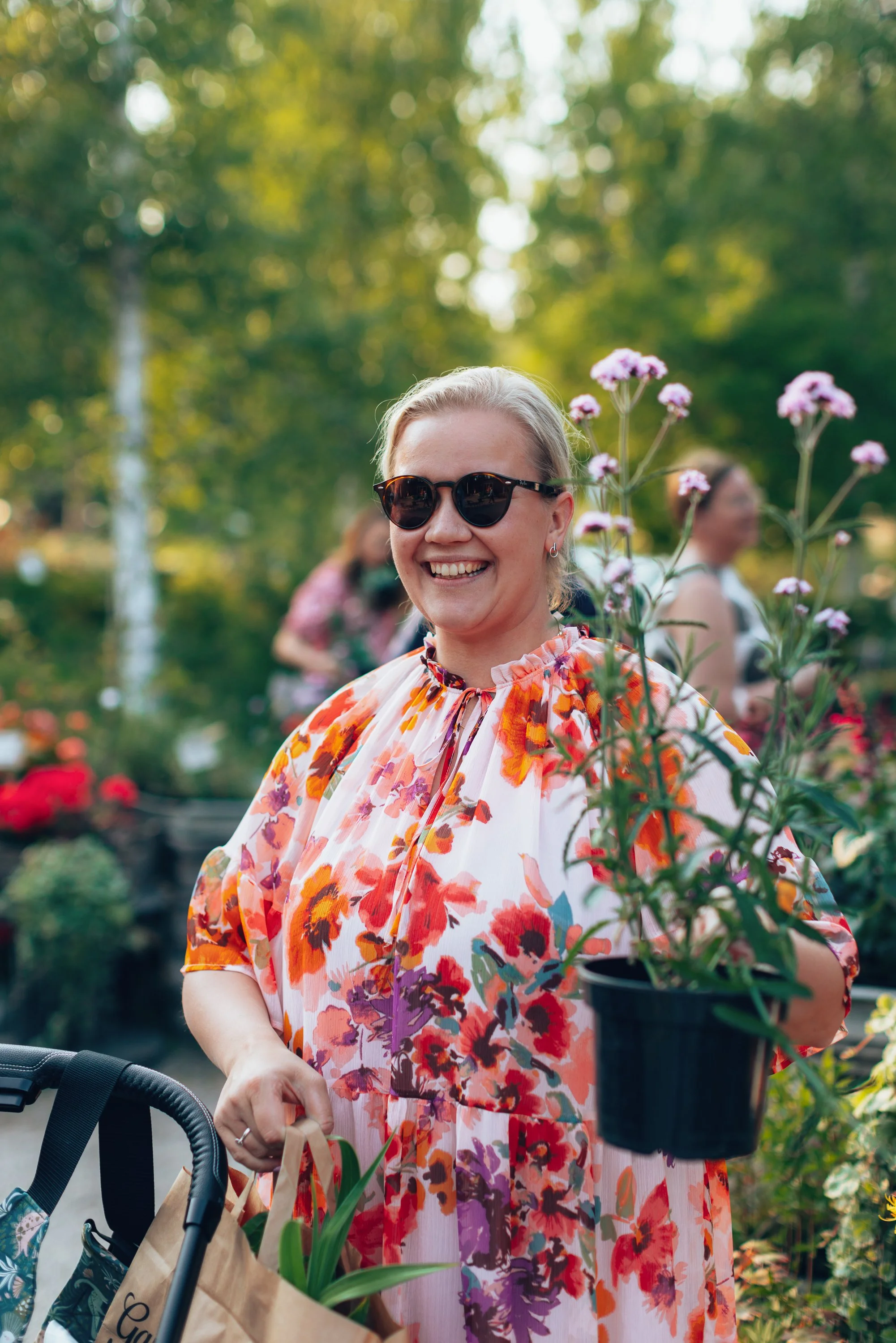 A woman smiling while holding a potted flower in a garden with other people in the background.