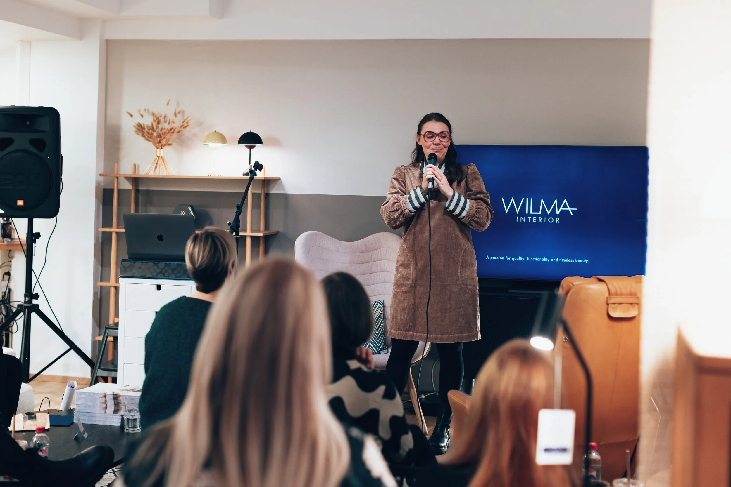 A woman in a brown coat and glasses speaks into a microphone in front of seated audience members in a room with a large screen that reads 'WILMA INTERIOR'.