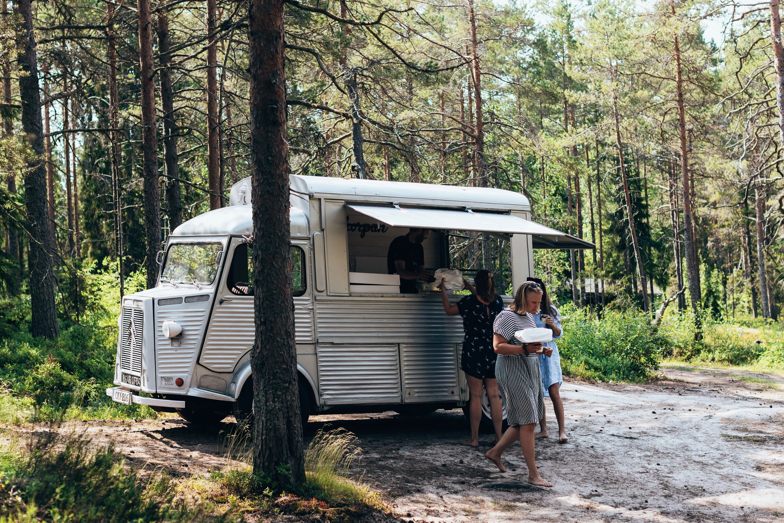 People enjoying food and drinks at a food truck in a forested outdoor area with tall trees and greenery.