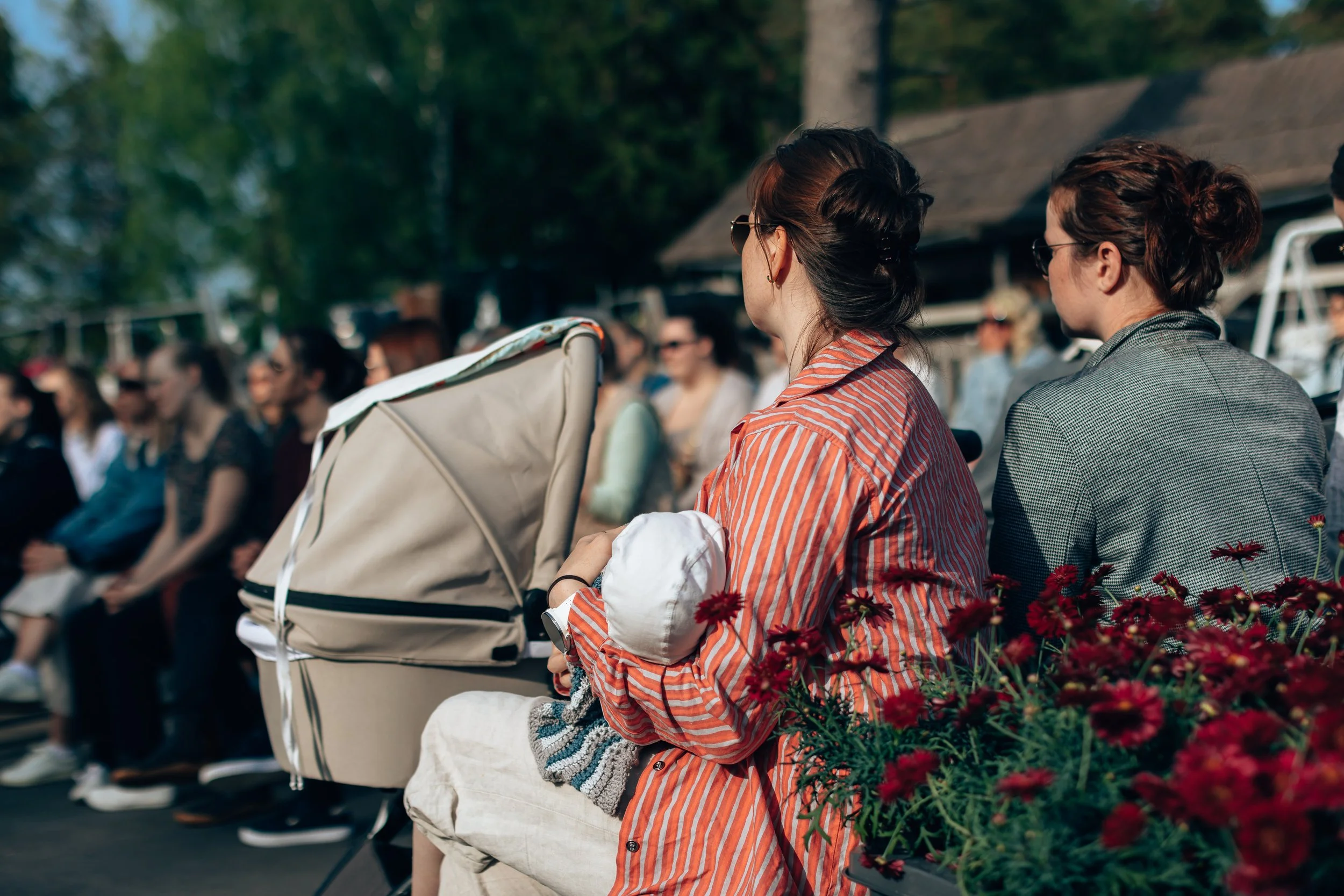 Women sitting outdoors at an event with a crowd in the background, some women are wearing sunglasses and striped shirts, one woman is holding a baby and a stroller, with red flowers nearby.