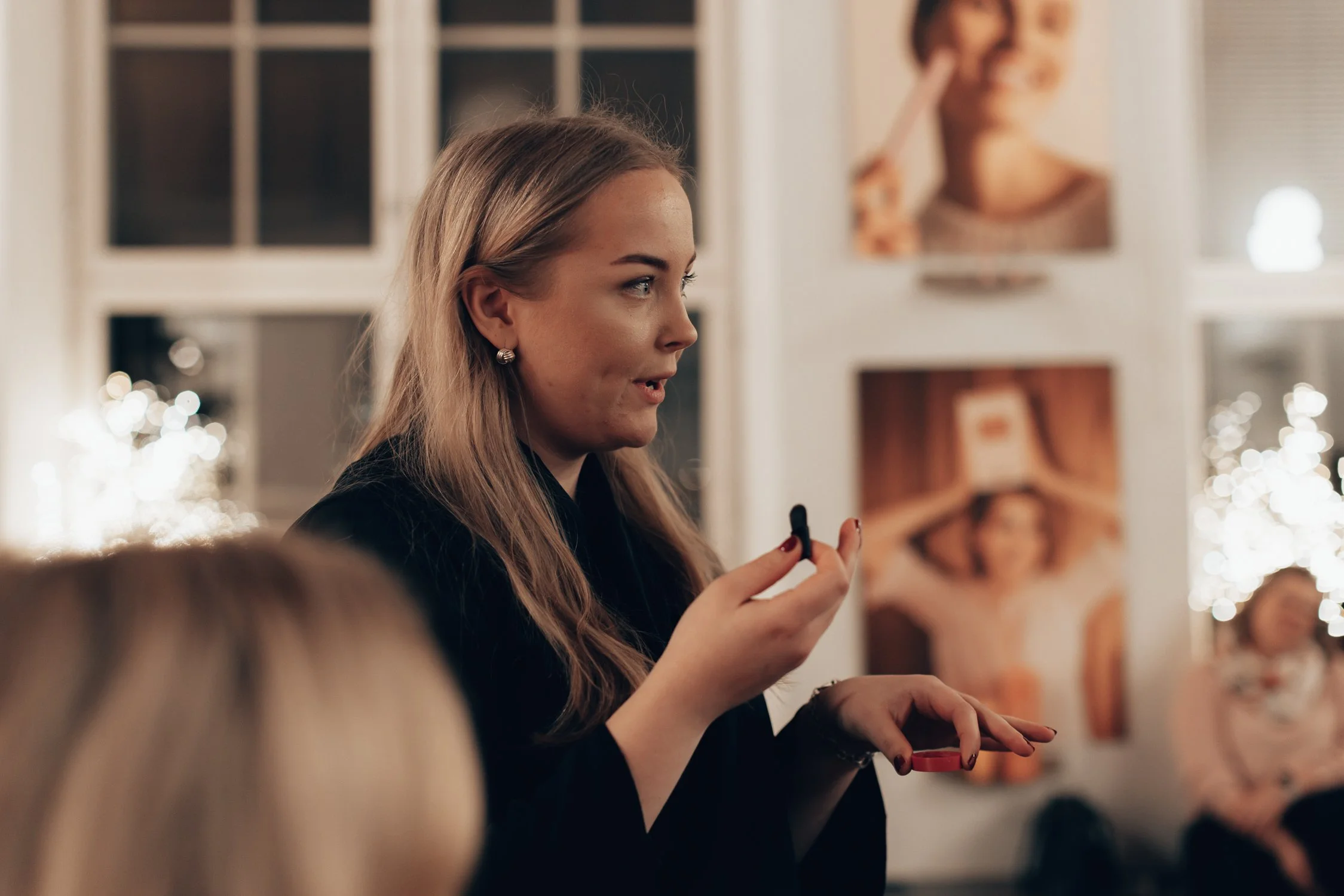 A woman with long blonde hair speaking, holding a microphone, indoors with blurred portraits on the wall in the background.