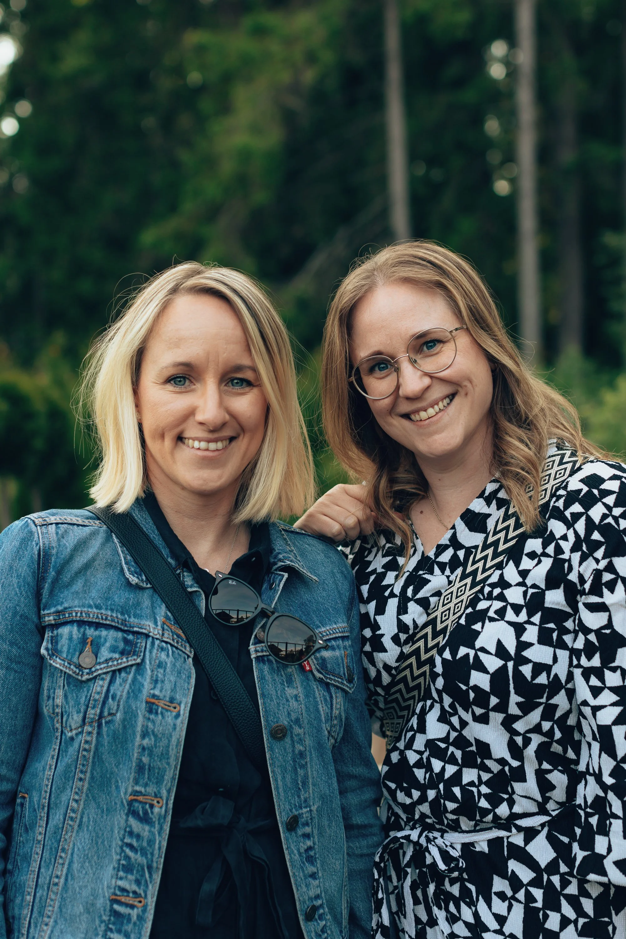 Two women smiling outdoors with trees in the background, one wearing a denim jacket and sunglasses, the other wearing glasses and a black and white patterned shirt.