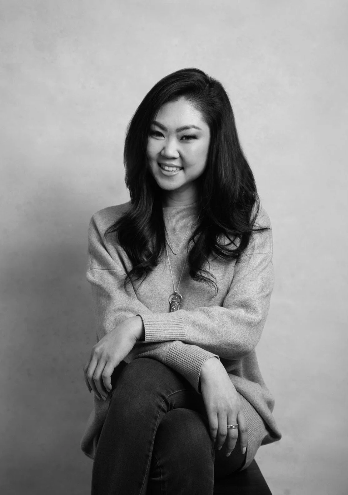 Black and white portrait of a young woman with long dark hair, smiling, wearing a sweater and necklace, sitting with crossed legs against a plain background.